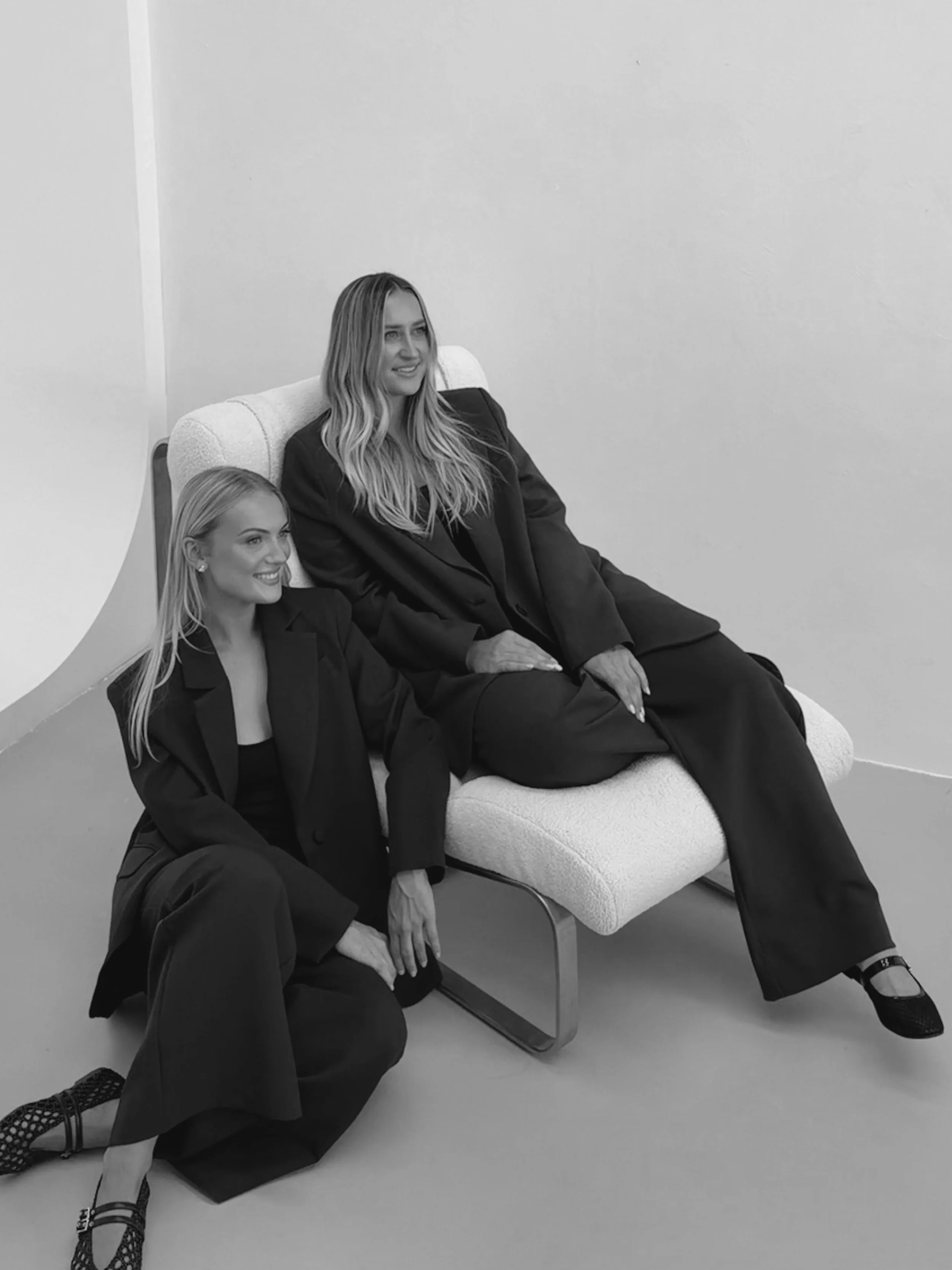 Two women dressed in black suits, one sitting on a couch and the other kneeling on the floor beside her, both smiling in a minimalist white room.