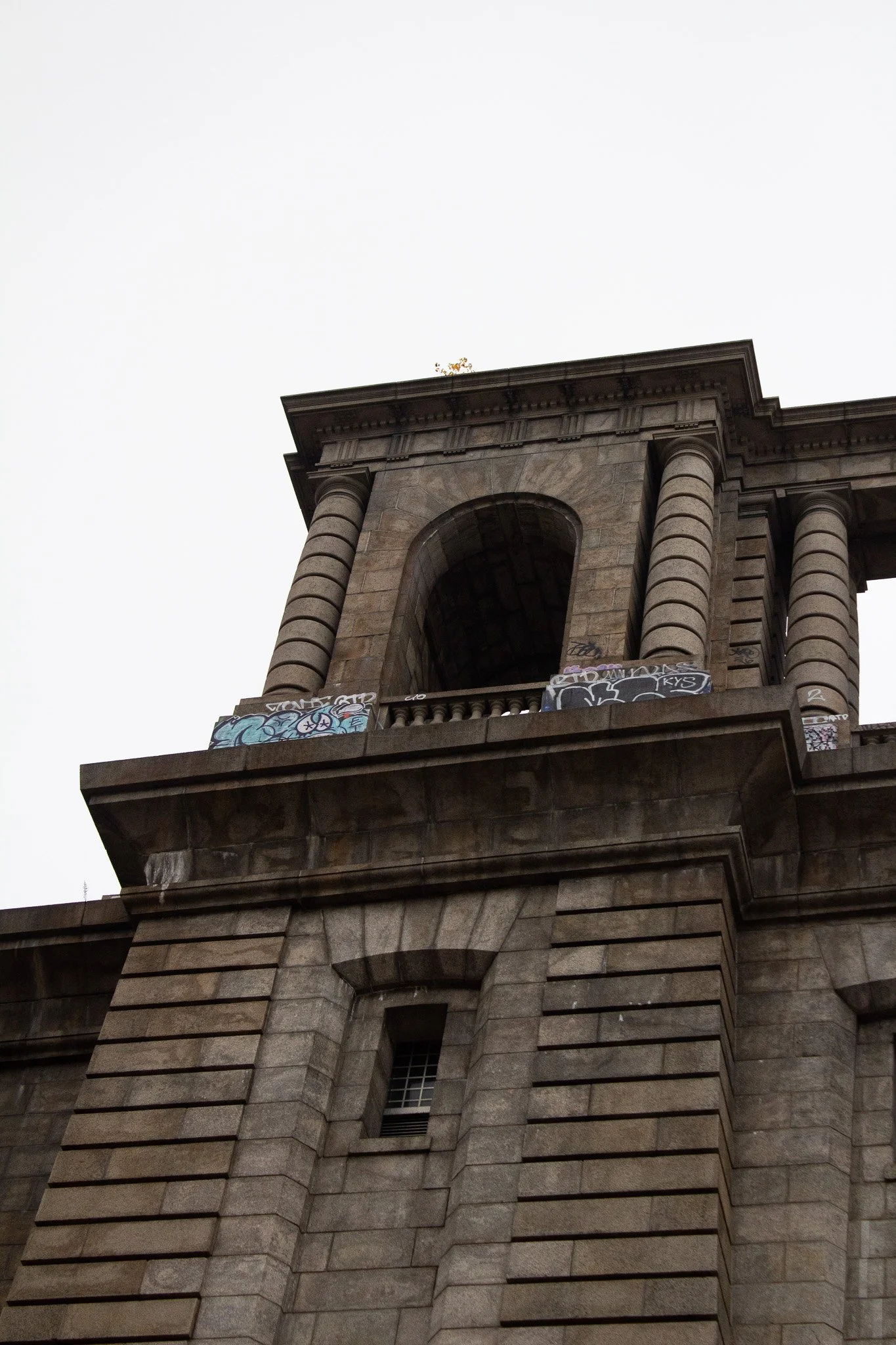 Vertical close-up view of a historic stone building or bridge with columns and an arched window, some graffiti on the ledge, against a gray sky.