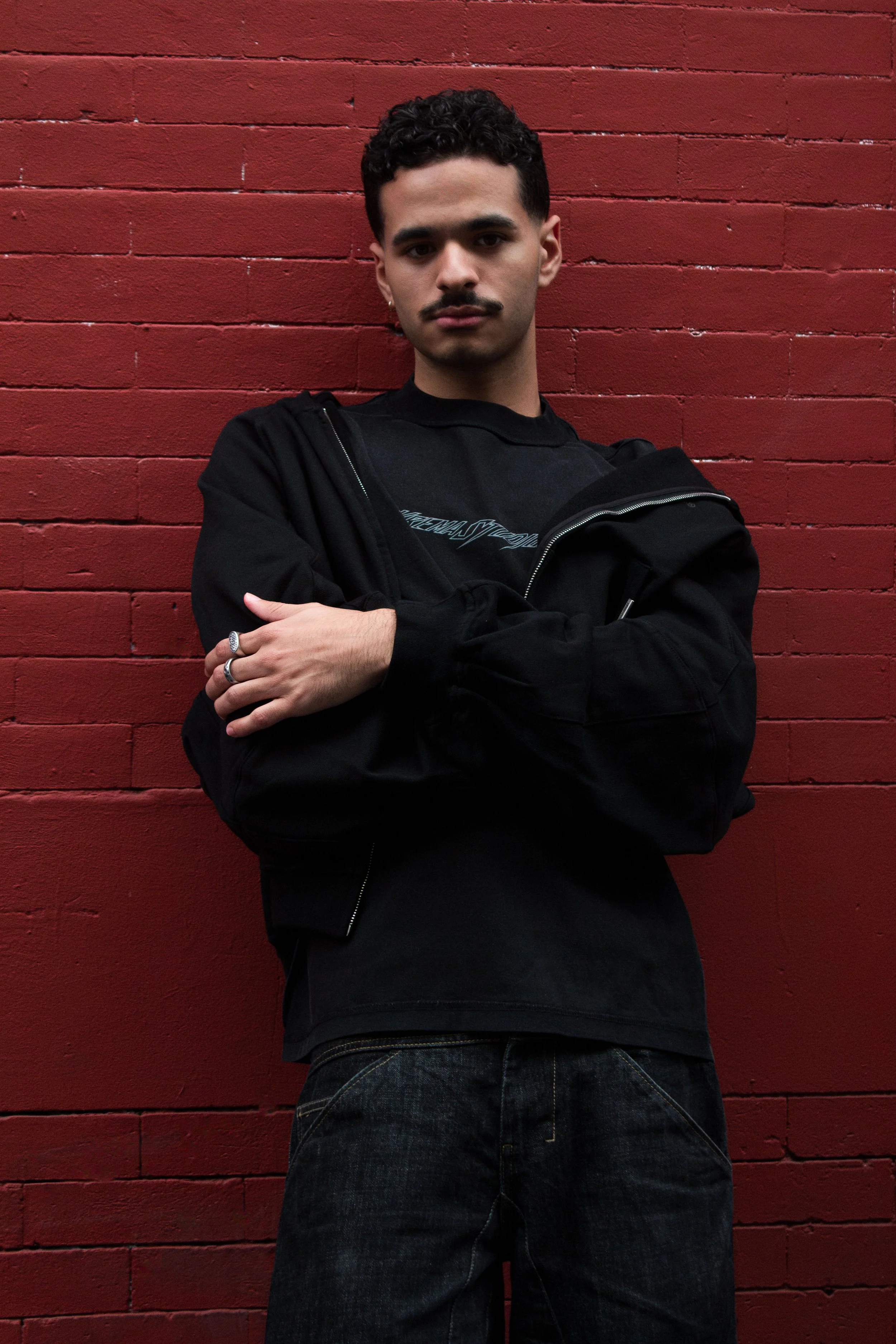 Young man with curly hair, mustache, and earrings stands against a red brick wall, crossing his arms, wearing a black jacket over a black graphic t-shirt and black jeans.