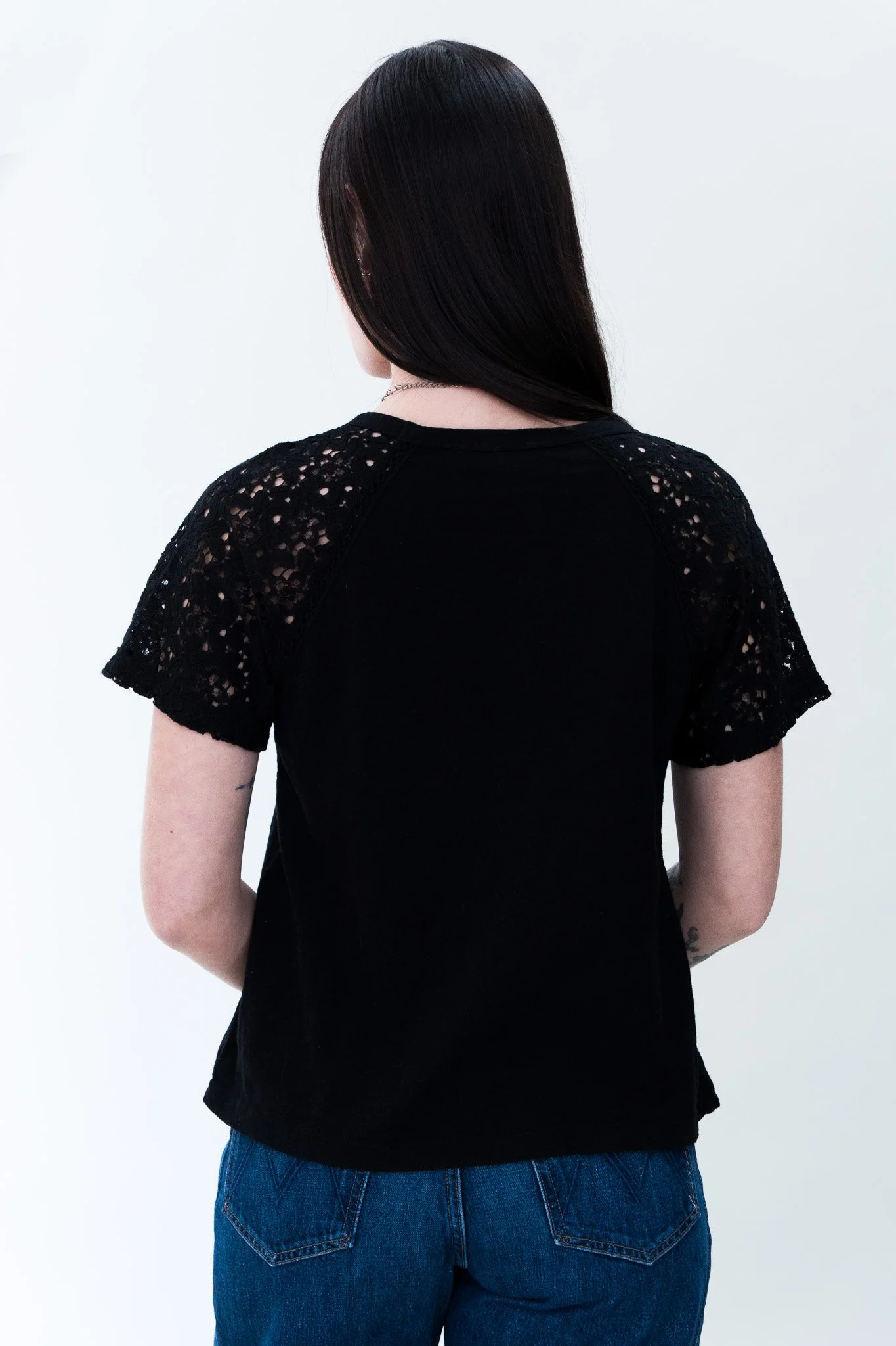 Back view of a person with long, straight dark hair wearing a black top with lace sleeves and blue jeans against a plain white background.