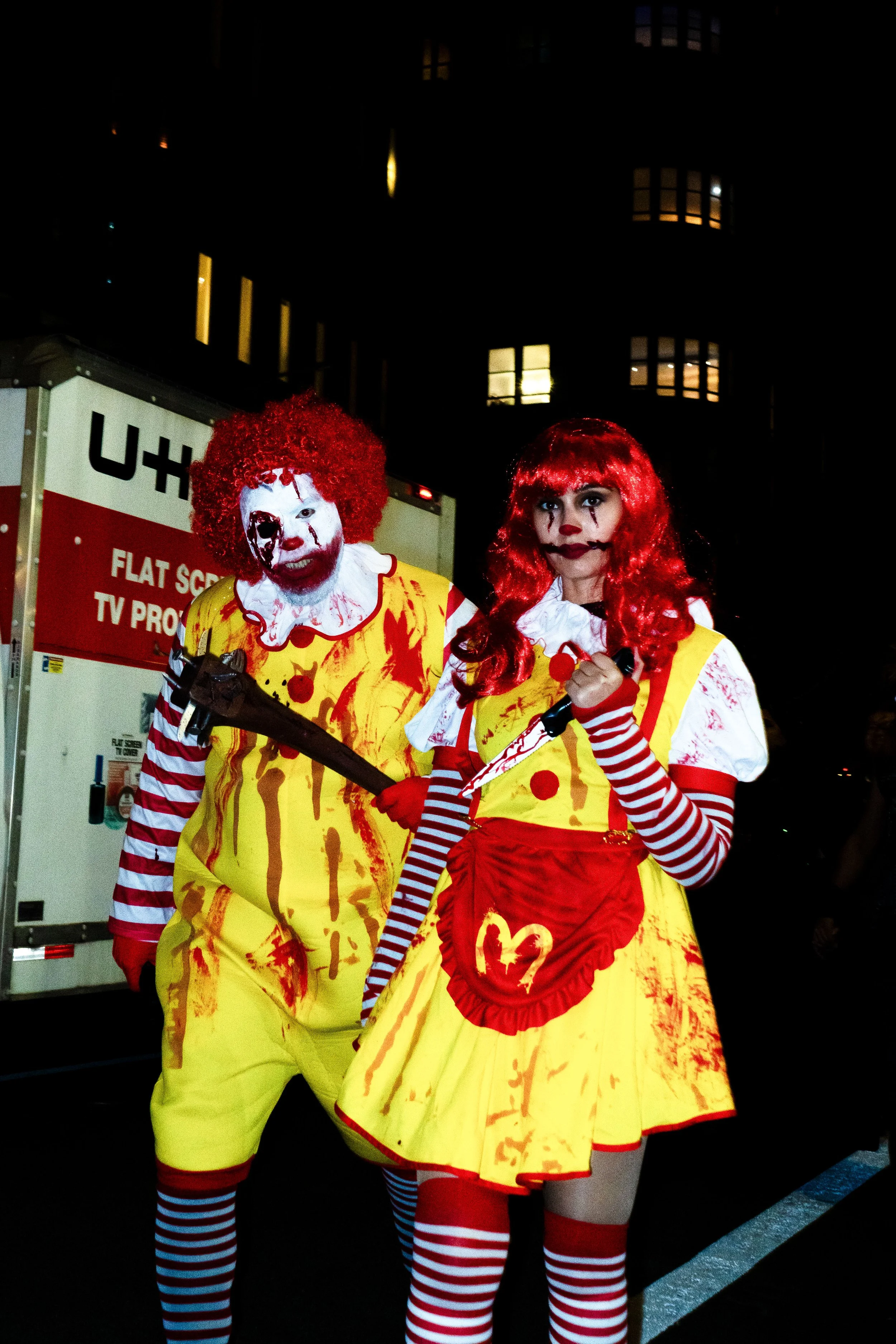 People dressed as creepy clowns in blood-stained costumes, with red wigs and striped stockings, standing outdoors at night.