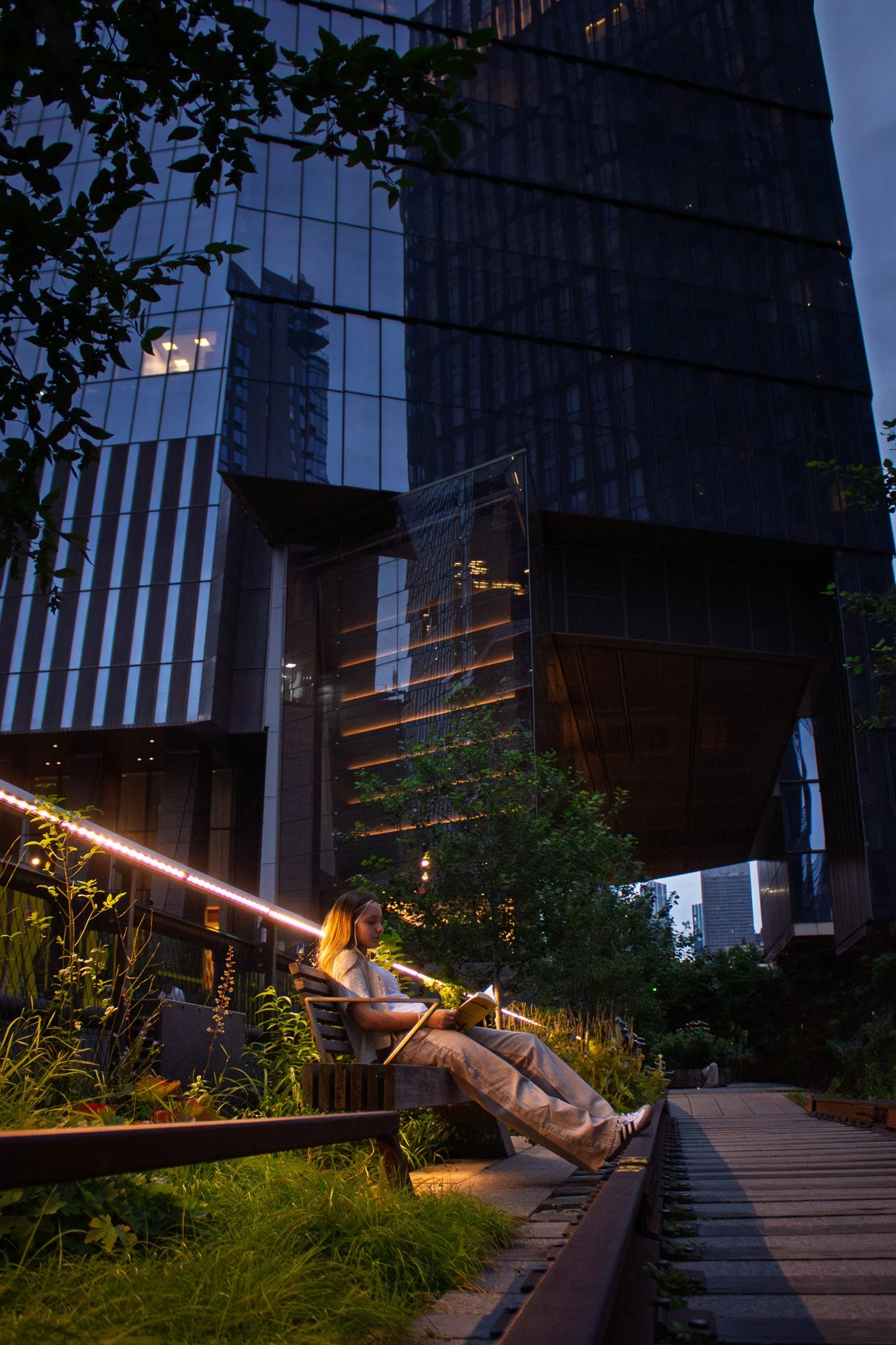 A woman sitting on a bench in a green area reading a book, with modern glass skyscrapers reflected in the windows behind her, illuminated by evening light.