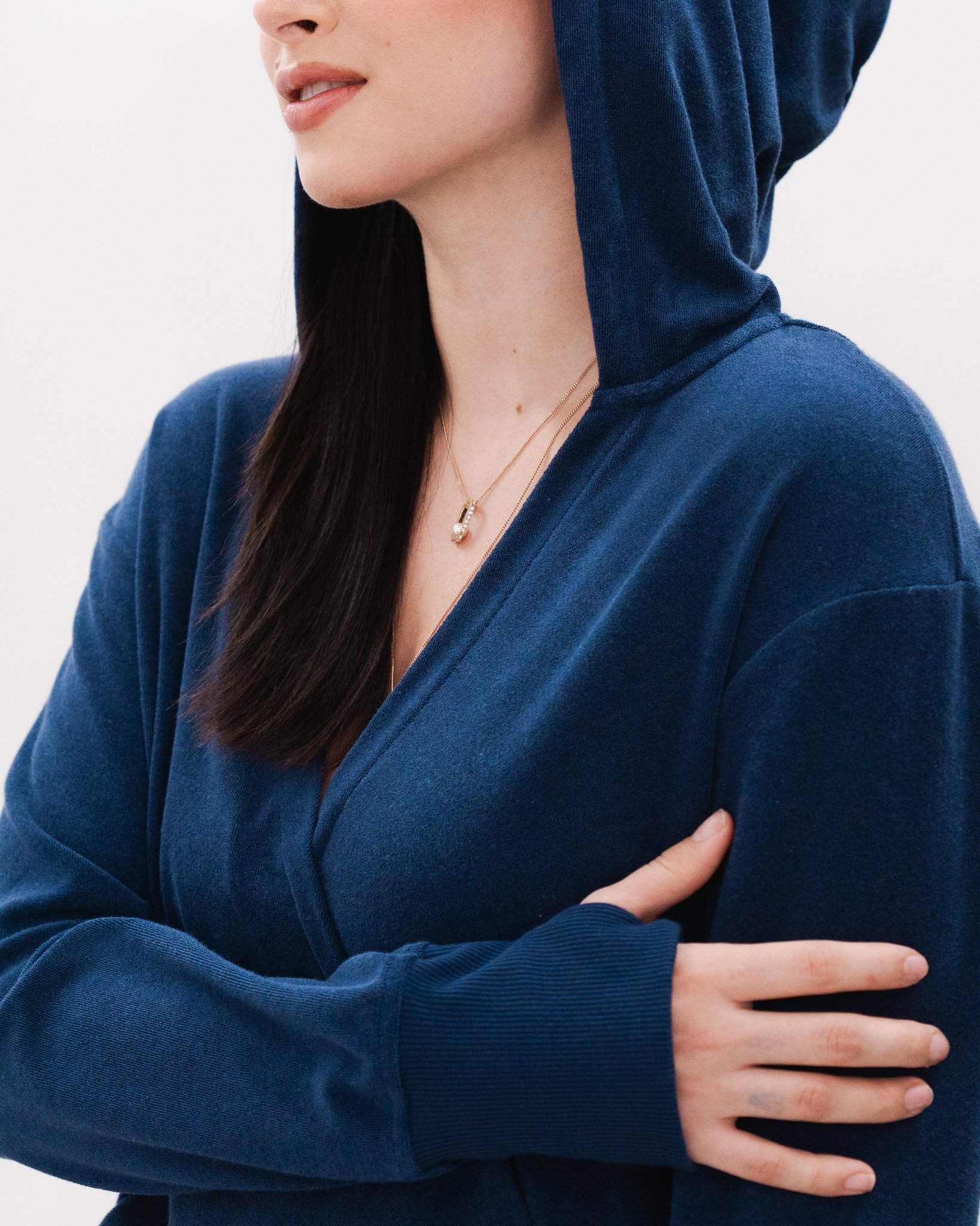 A person wearing a navy blue hoodie, with their arm crossed over their chest, showing a gold necklace with a pendant.