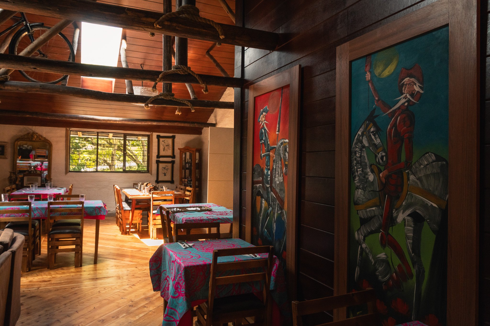 Inside a cozy restaurant with wooden walls and ceiling, decorated with vibrant paintings of figures in traditional attire. Tables are covered with colorful, patterned tablecloths, and a large window allows natural light to illuminate the space.