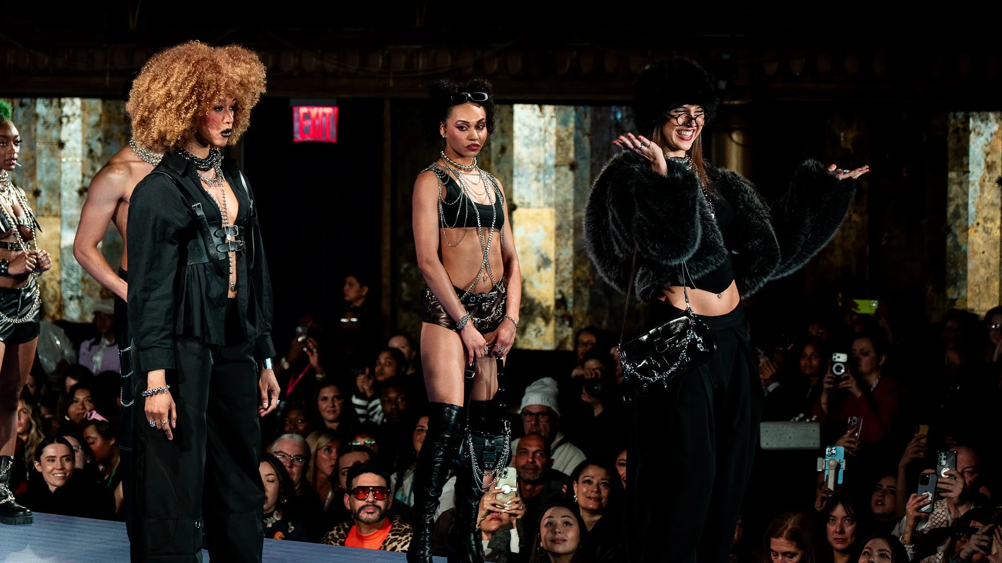 Group of diverse women modeling fashionable outfits on a runway with a crowd in the background.