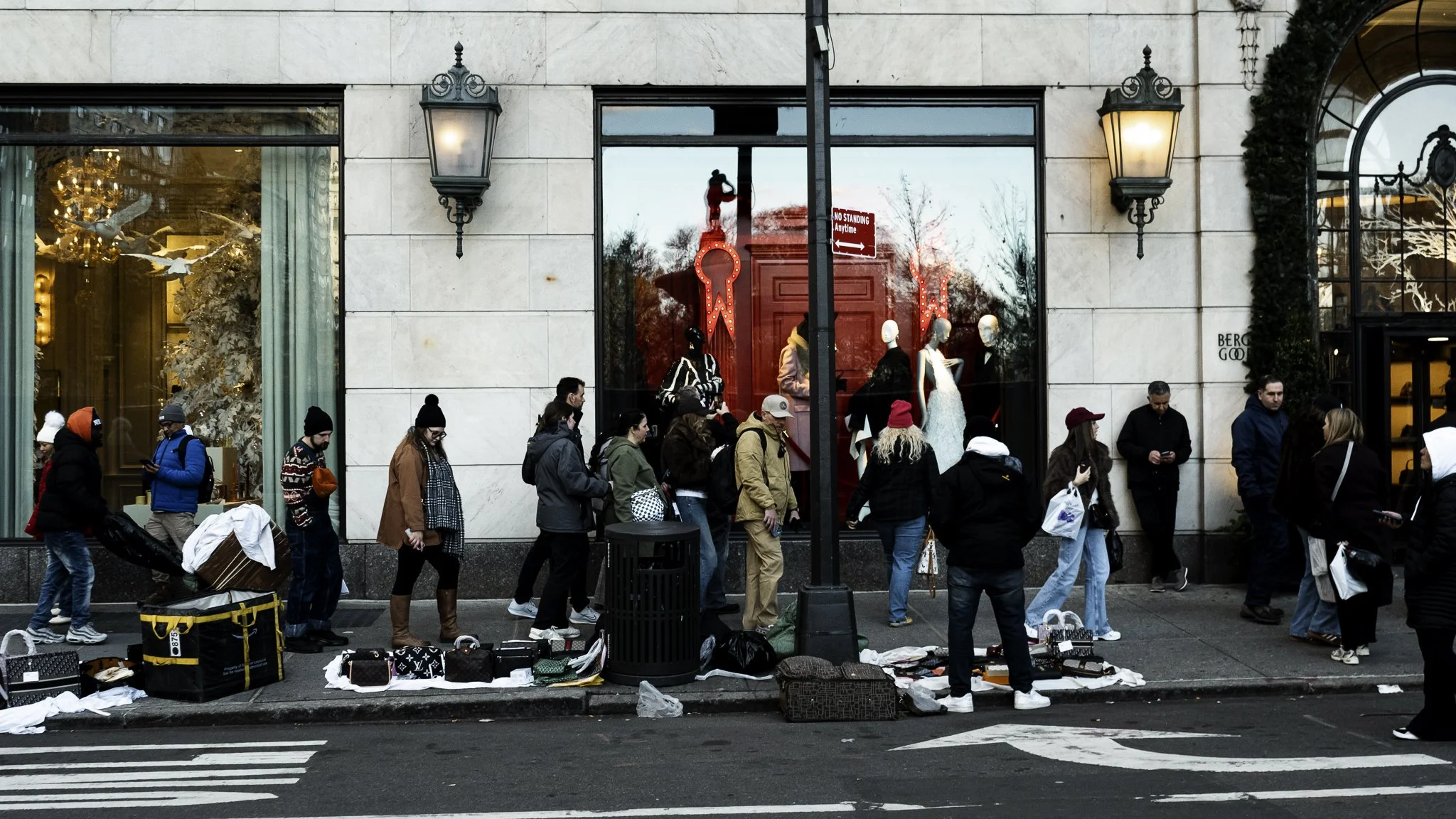 People standing and shopping outside a storefront with mannequins in display windows, bags and luggage on the sidewalk, and street crossing lines in the foreground