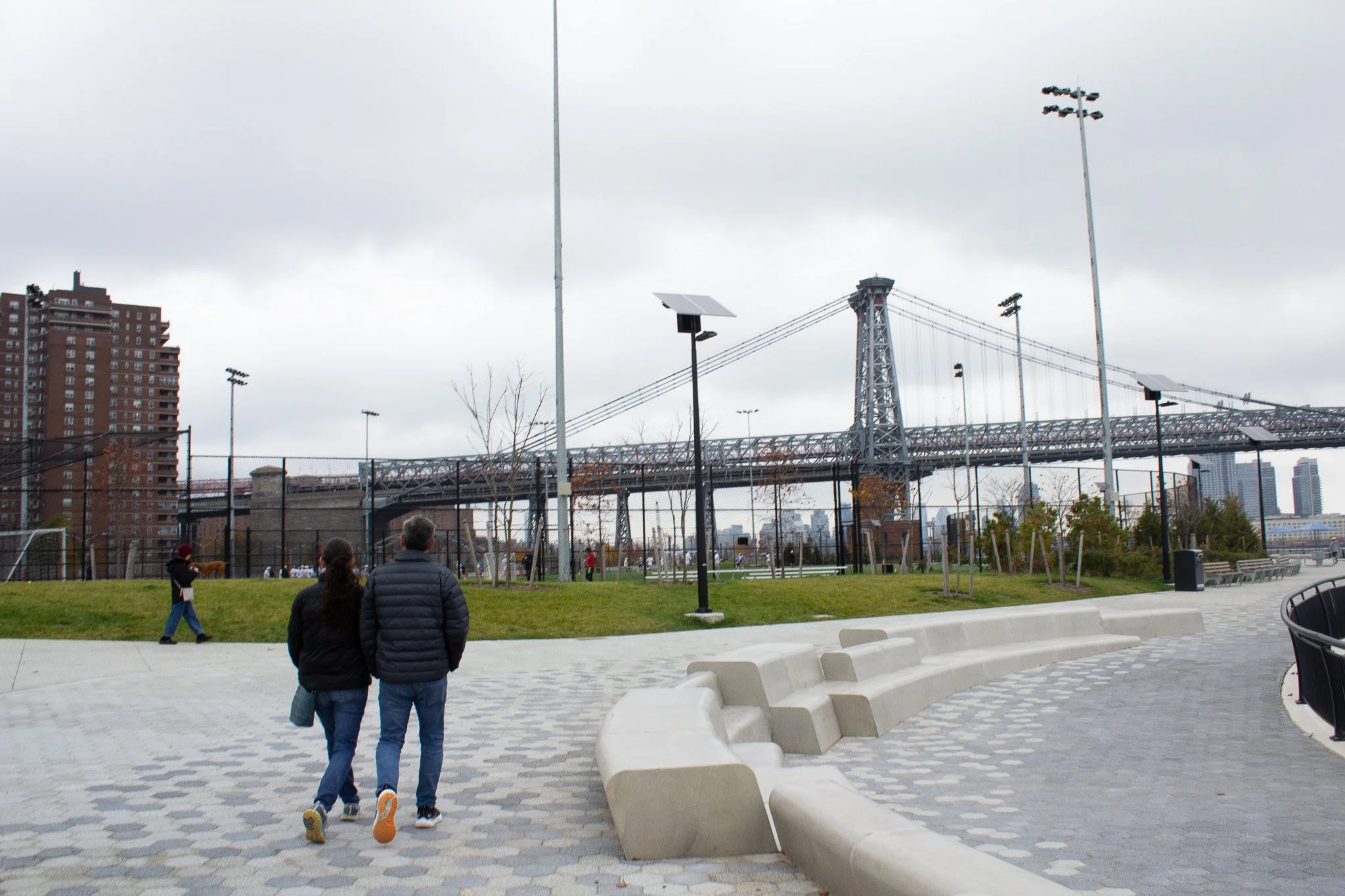 A park with a pathway, modern concrete seating, and children playing basketball. In the background, there is a bridge and residential buildings under a cloudy sky.