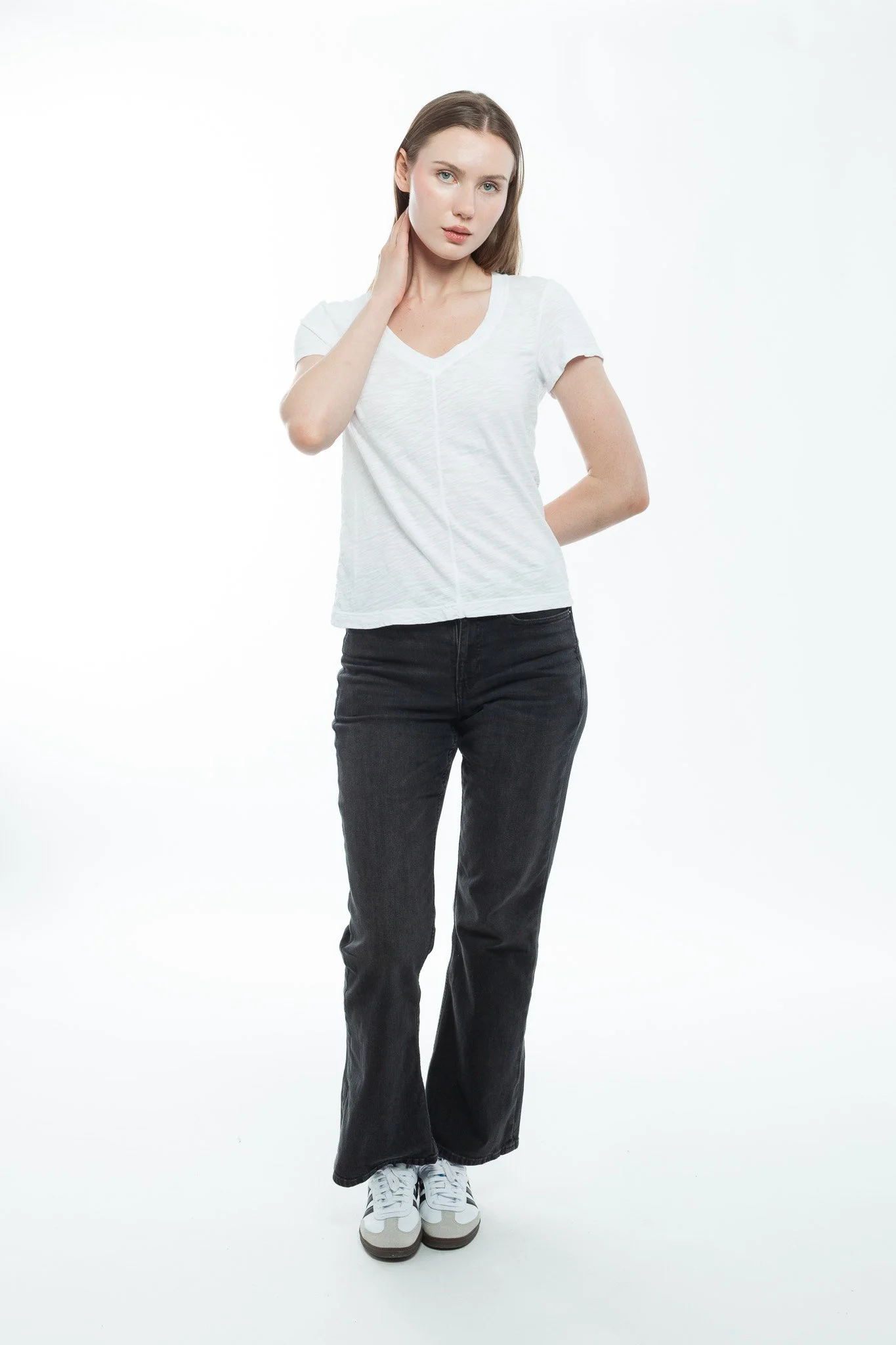 A young woman with long brown hair wearing a white t-shirt, black jeans, and white sneakers, standing against a plain white background, touching her neck with her right hand.