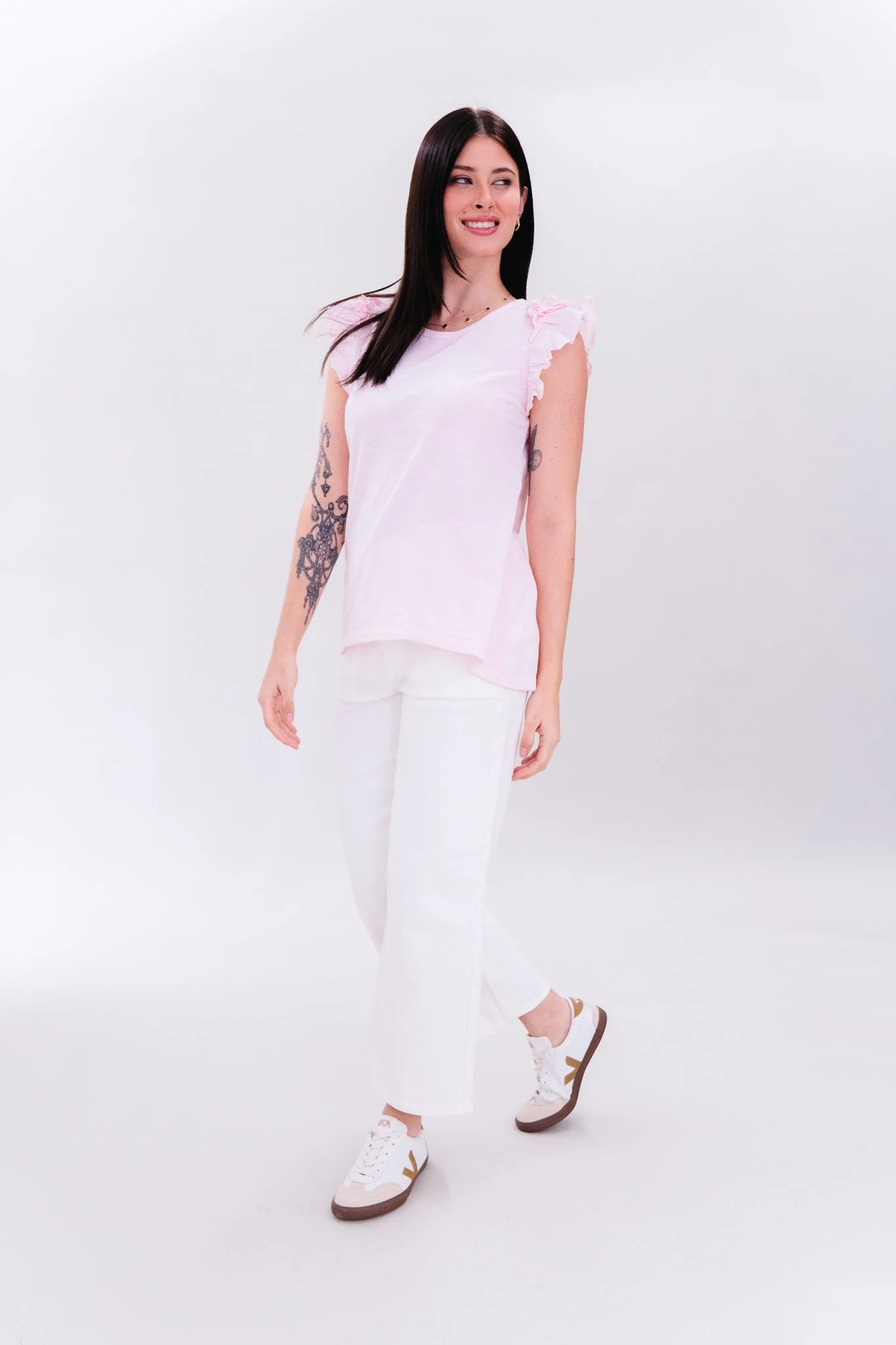 A woman with black hair, wearing a light pink sleeveless top with ruffled shoulders, white pants, and white sneakers with brown accents, standing against a plain white background, smiling and looking to the side.