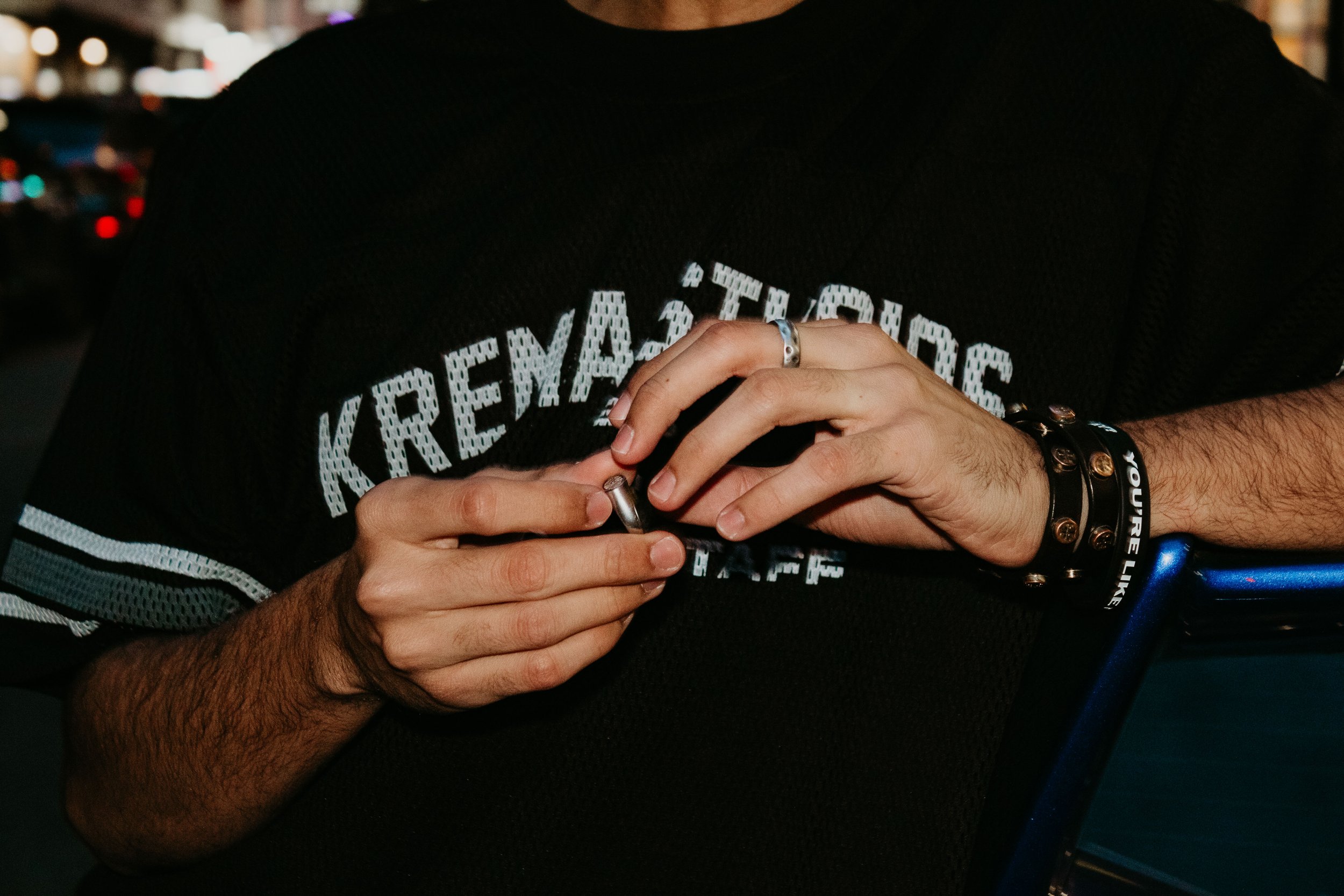 A person wearing a black sports jersey with white lettering, sitting outdoors at night, holding a small item in their hands, wearing a ring on the right hand and a black wristband on the left wrist.