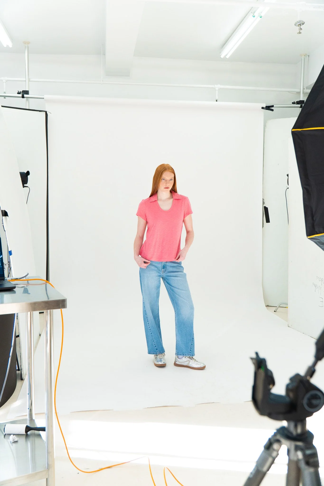 A young woman with red hair and a pink t-shirt stands in front of a white backdrop in a photography studio, surrounded by studio lighting and equipment.