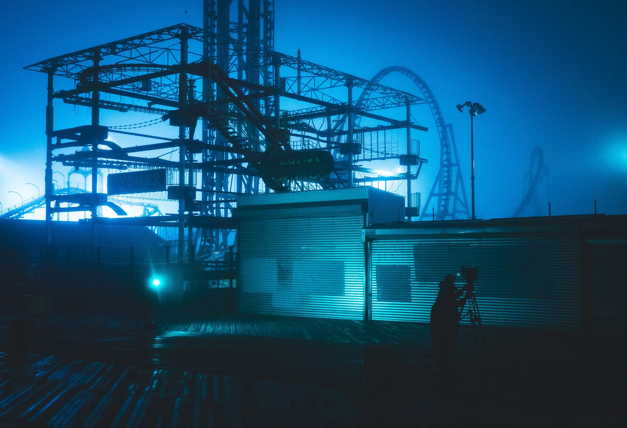 Dark amusement park ride structures illuminated by blue light at night, with silhouetted people nearby.