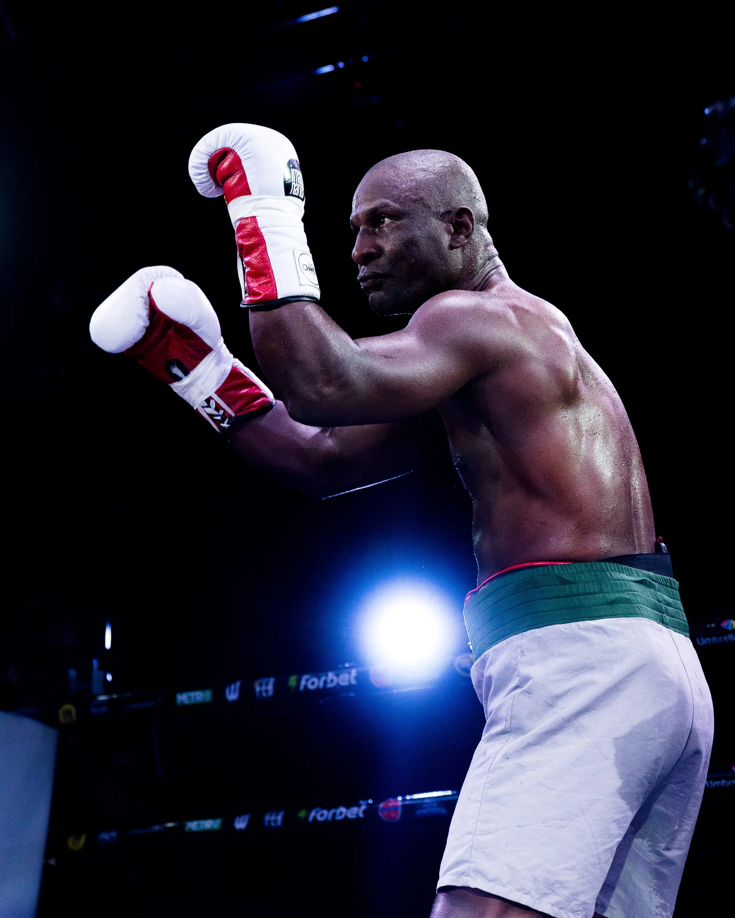 Boxing fighter with raised fists, sweating, wearing red and white gloves, white shorts with green trim, under bright lights in a dark arena.