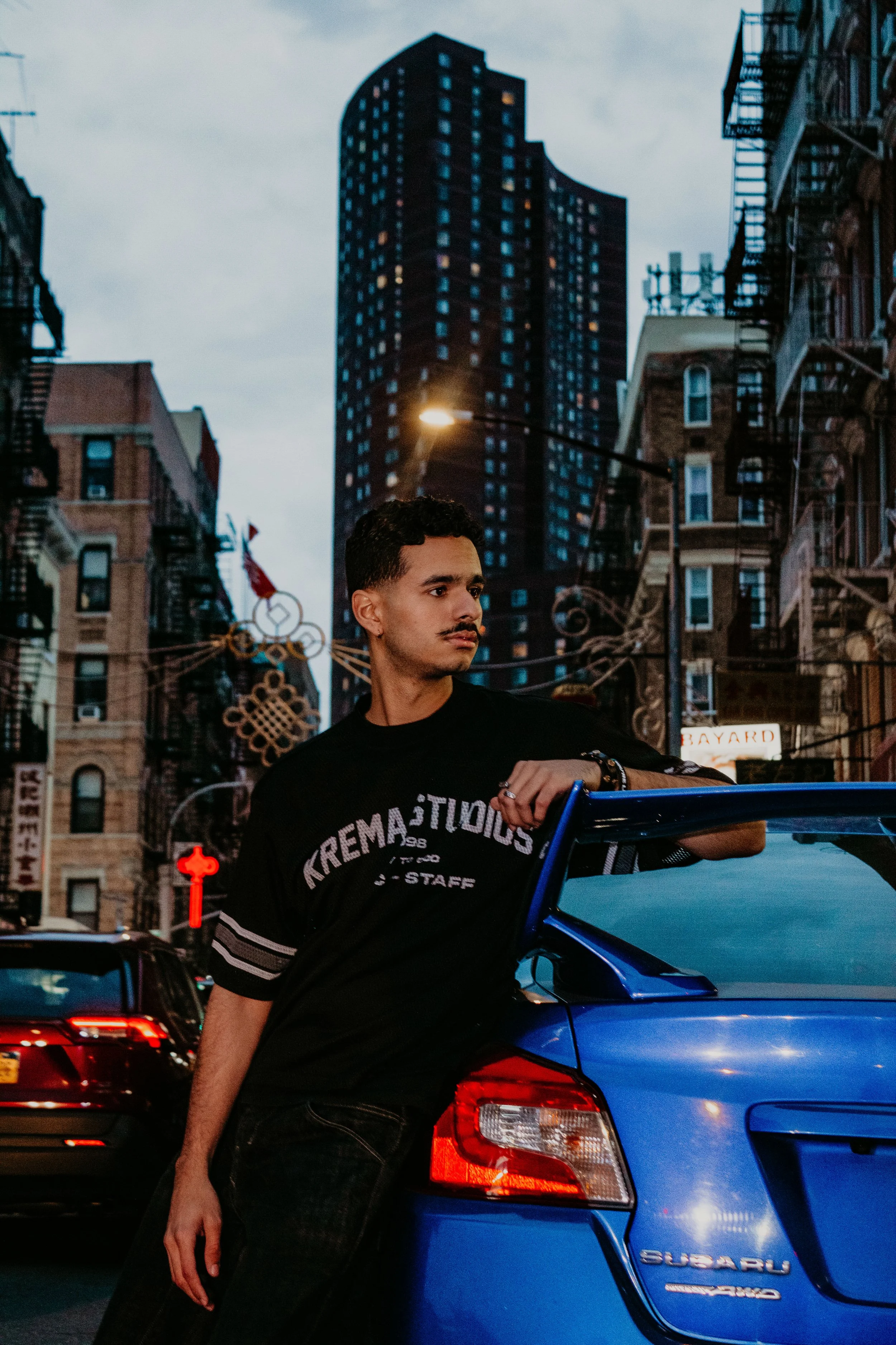 A young man with short curly hair and a mustache leaning against a blue Subaru car on a city street during dusk, with tall buildings and streetlights in the background.