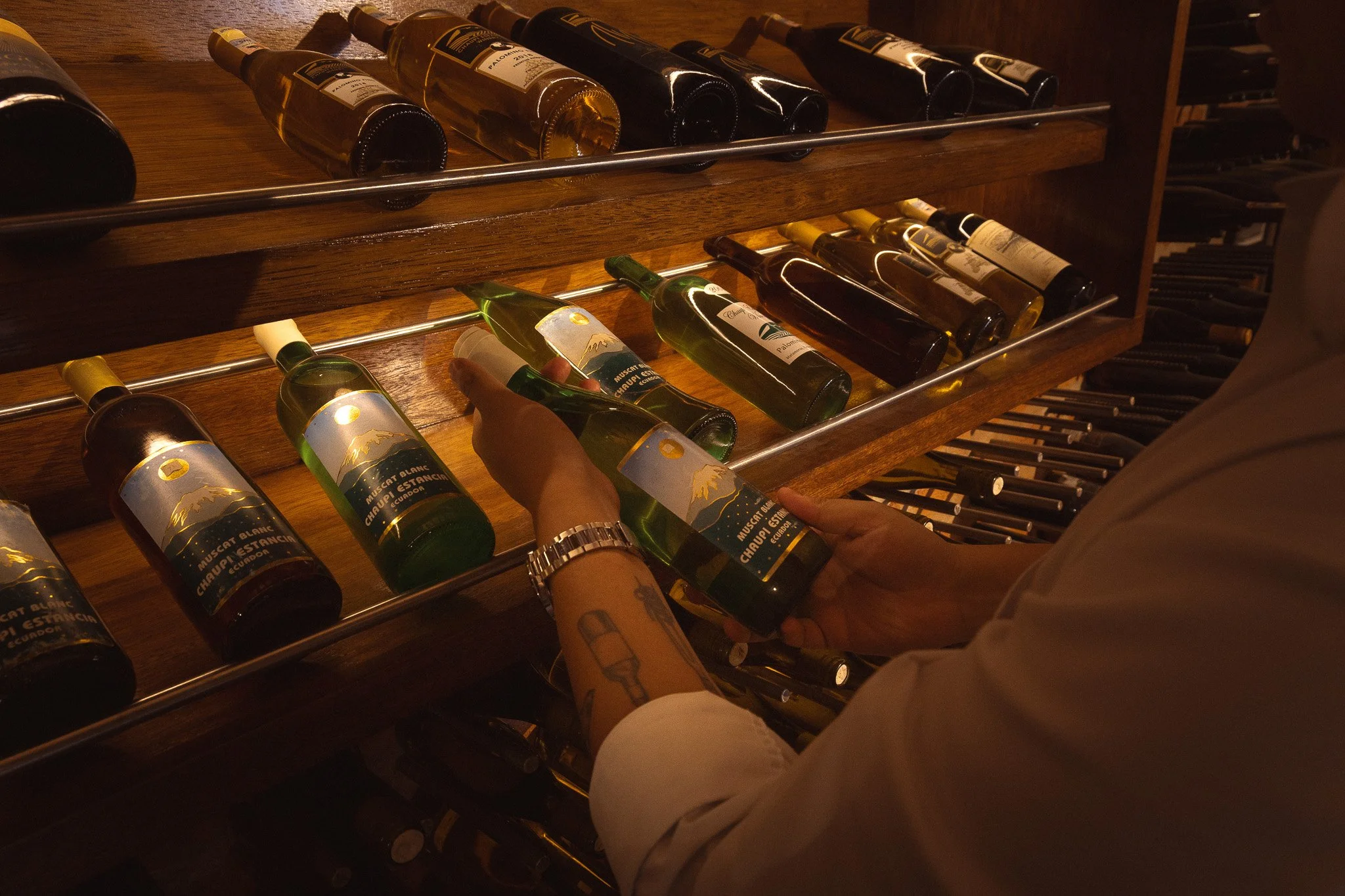 Person with tattoos and a watch organizing bottles of wine on a wooden wine rack in a dimly lit room.