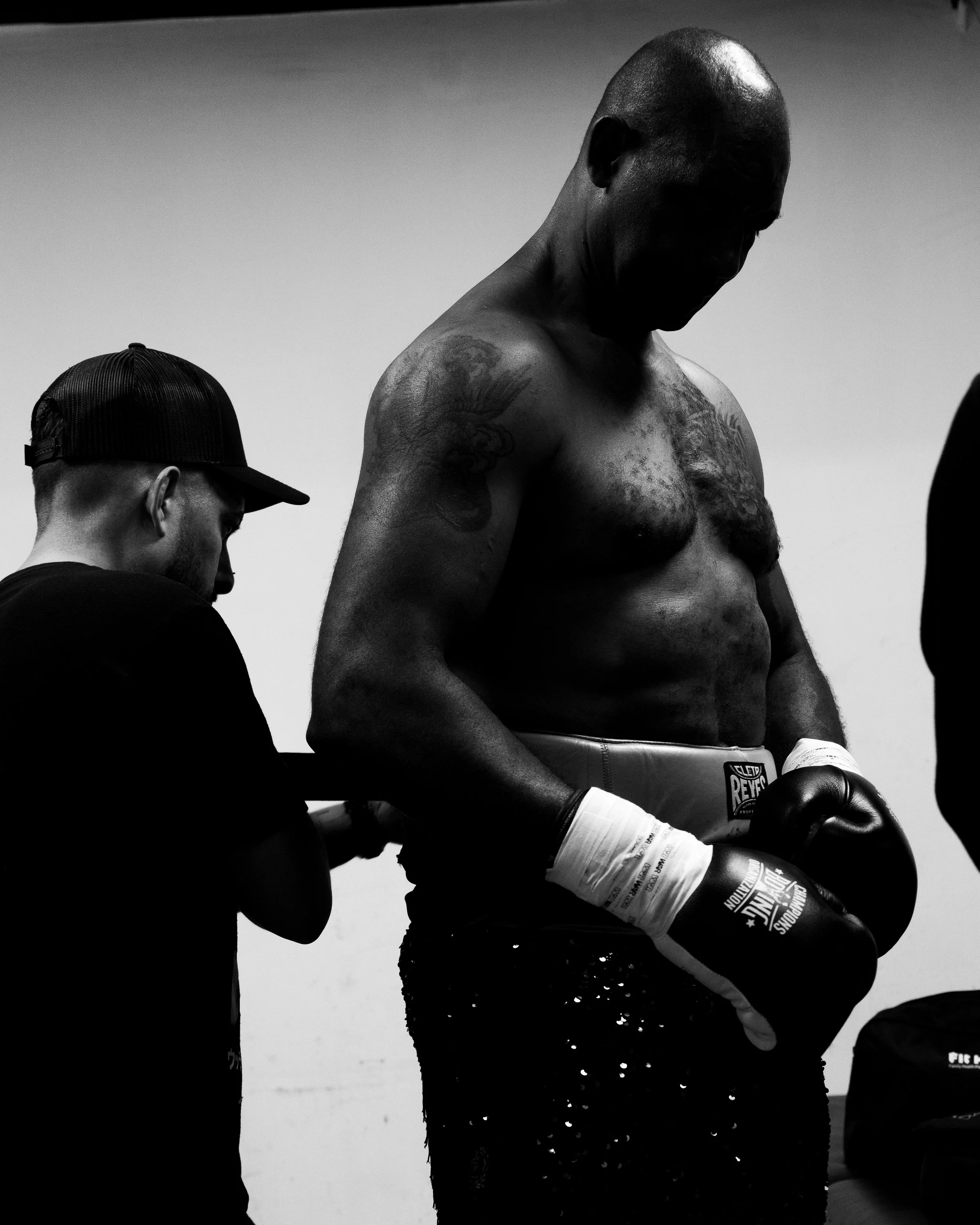 Black and white photo of a shirtless male boxer with tattoos, wearing boxing gloves and shorts, standing with other individuals around him in a gym or locker room setting.