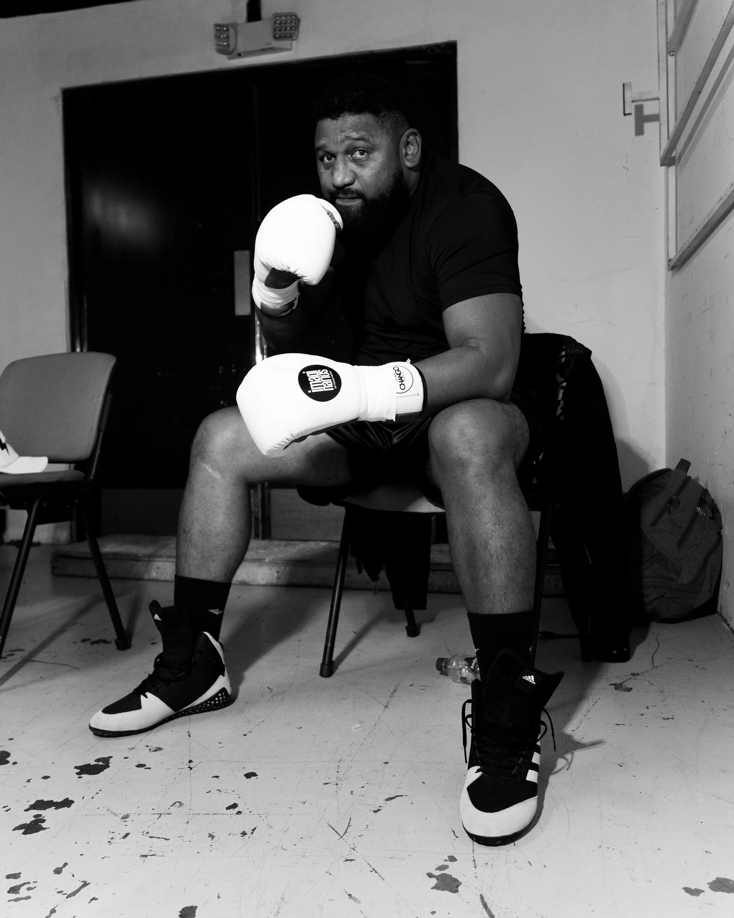 A man in boxing shorts and gloves sitting on a chair in a room with a marble floor. The man is wearing black and white boxing shoes and has a sports bottle and a backpack nearby.