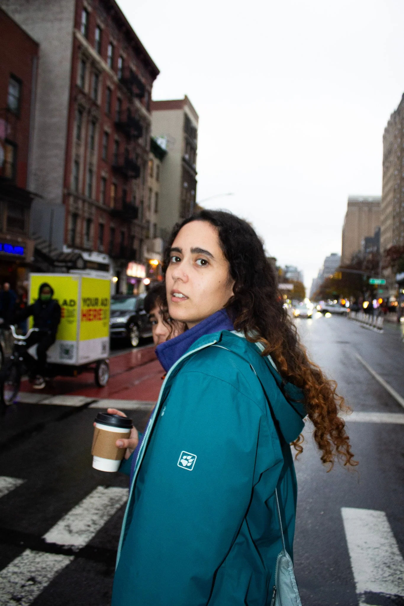 Young woman with curly hair holding a coffee cup, standing on a city street with buildings, cars, and a crosswalk, overcast weather.