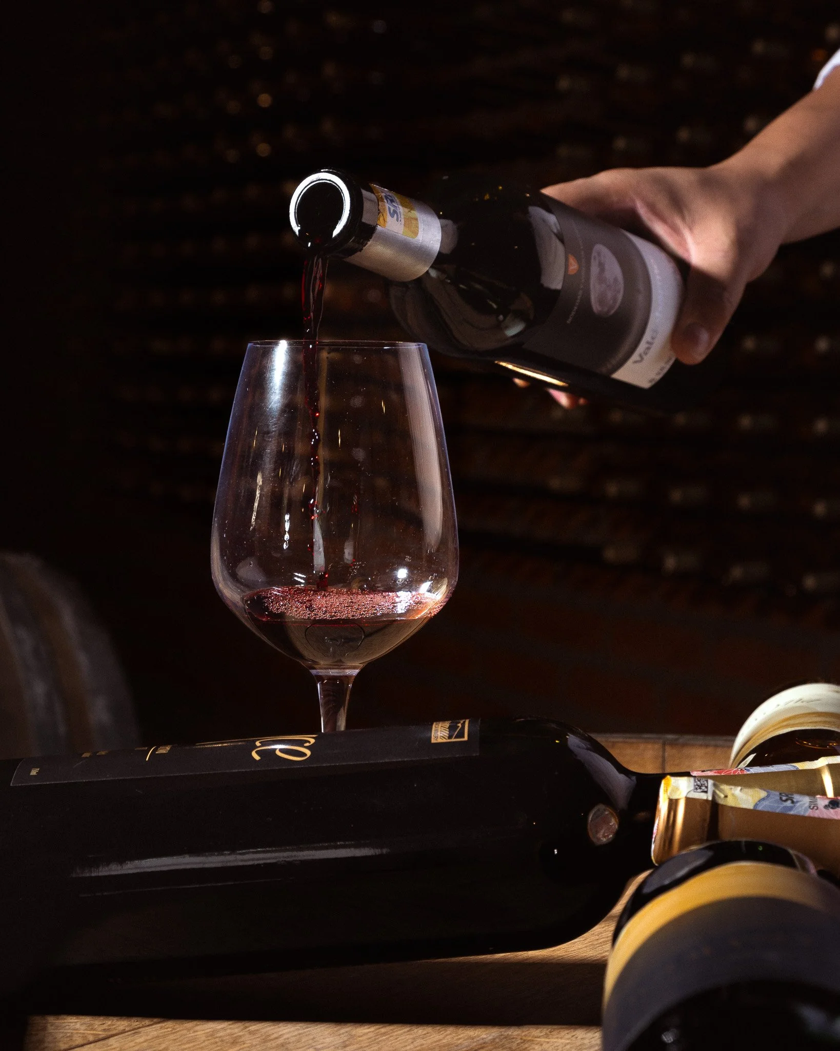 Person pouring red wine into a glass, with a wine bottle in their hand, on a dark wooden surface.