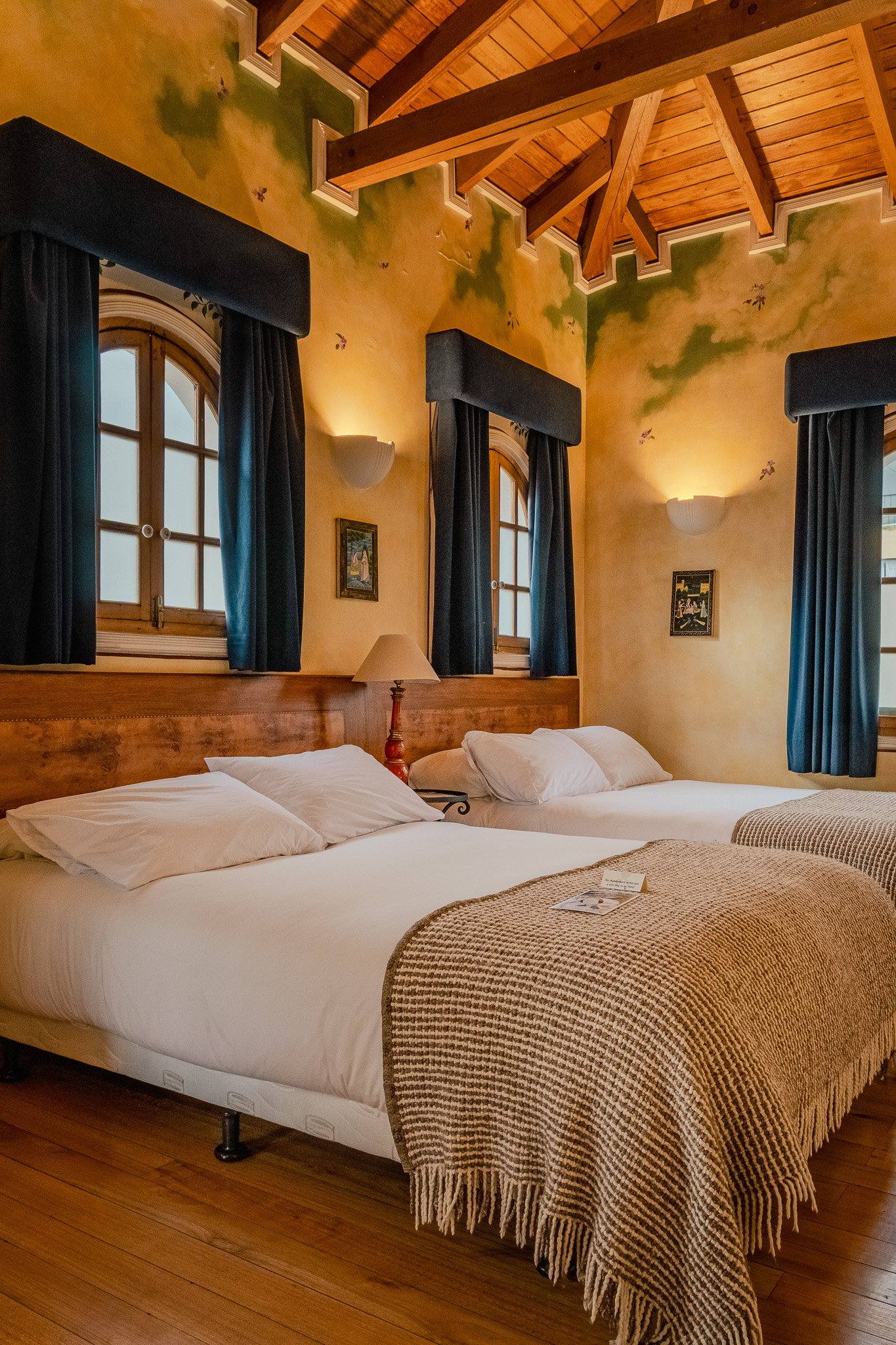 A cozy bedroom with two beds side by side, a blanket on one, and pillows. The wall behind features arched windows with dark curtains, wooden ceiling with exposed beams, and wall-mounted lights. There are small framed pictures on the wall.