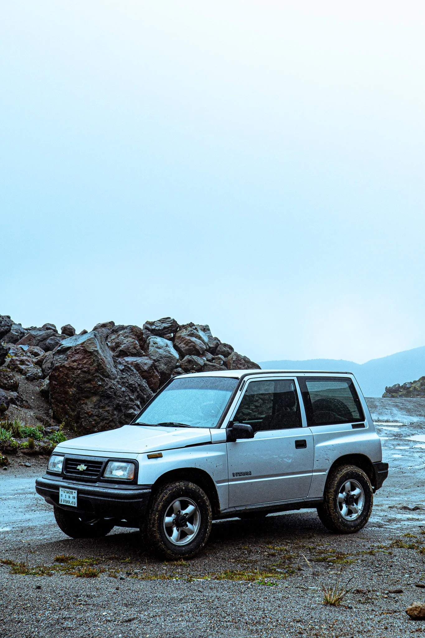 A small silver Chevrolet Vitara SUV parked on a gravel surface next to large rocks and a body of water, with mountainous terrain in the background under an overcast sky.