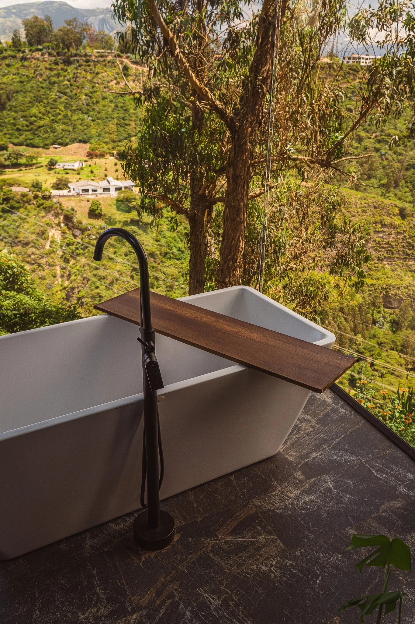 A view from a balcony with a white bathtub in the foreground, a black faucet, and a wooden tray resting on the edge. Behind, there are green trees and hills, with a large tree trunk next to the tub and mountains in the distance.