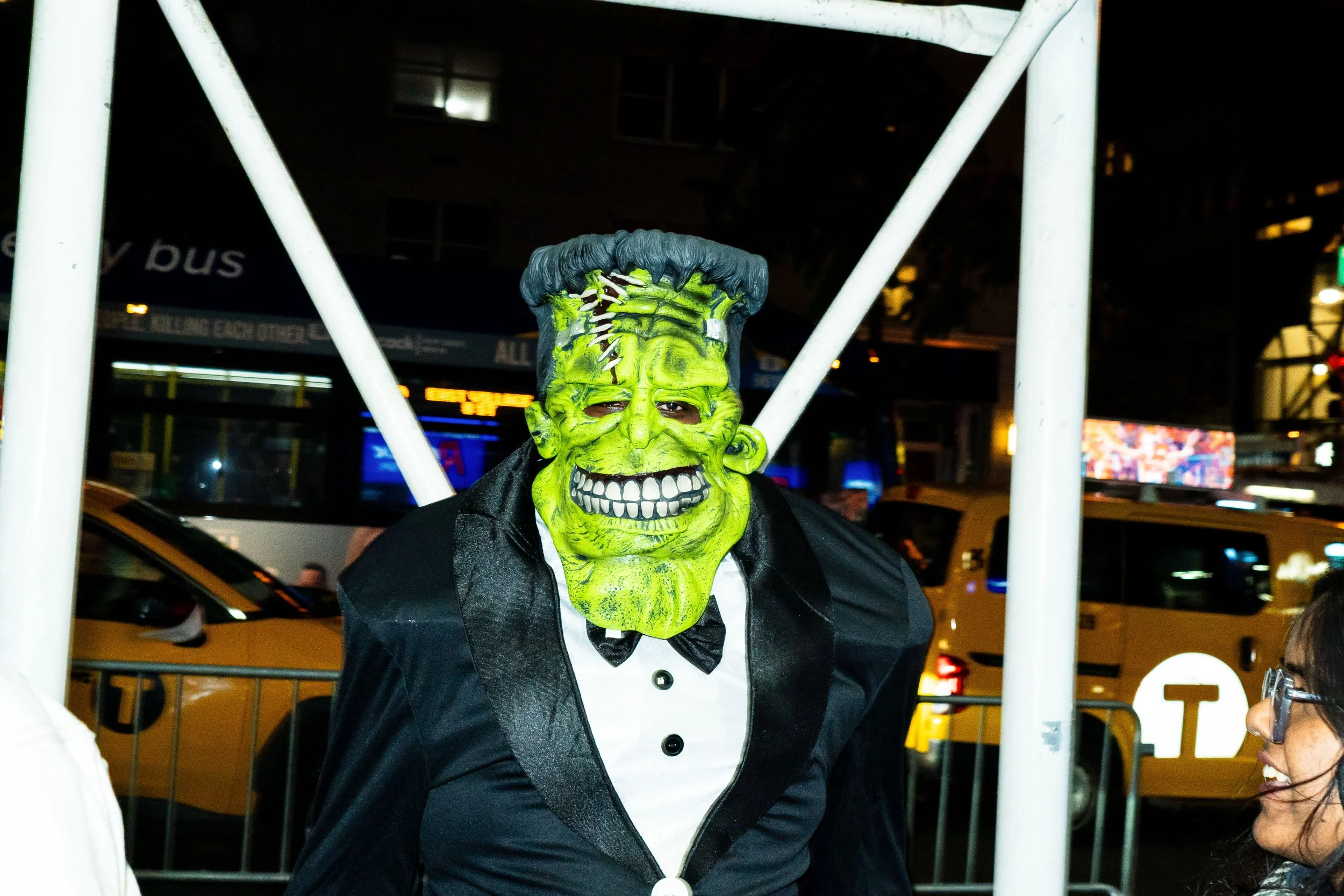 Person dressed in a Frankenstein monster costume with a green face, black hair, stitched forehead, wearing a tuxedo at night in a city street.