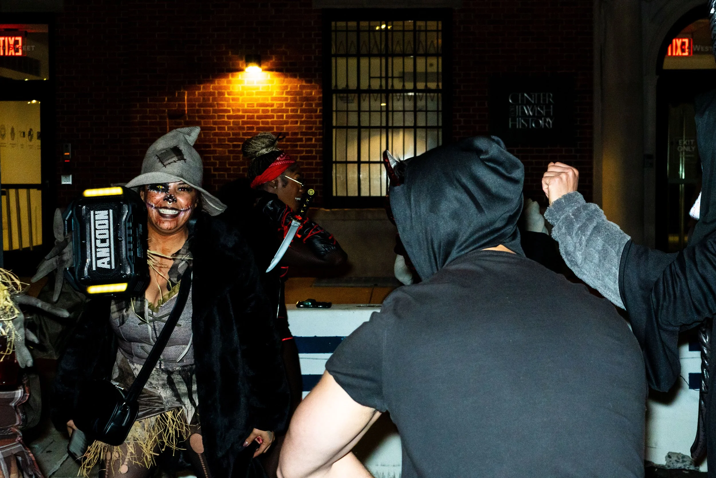 People dressed in Halloween costumes, including a witch and a spooky character, at a nighttime event outside a brick building.