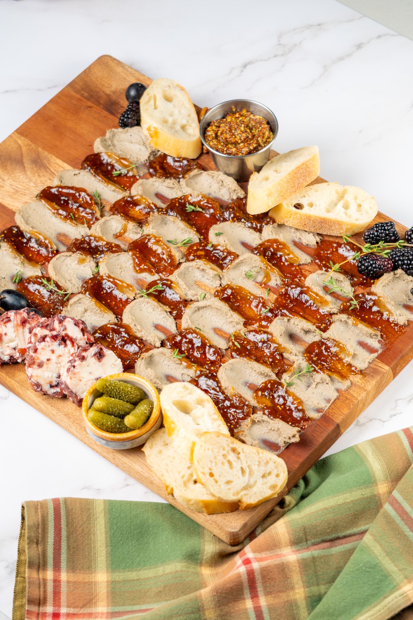 A wooden platter with sliced meat topped with barbecue sauce, sliced baguette bread, blackberries, a small bowl of mustard, green pickles, and other assorted bread slices on a marble surface, over a colorful cloth.