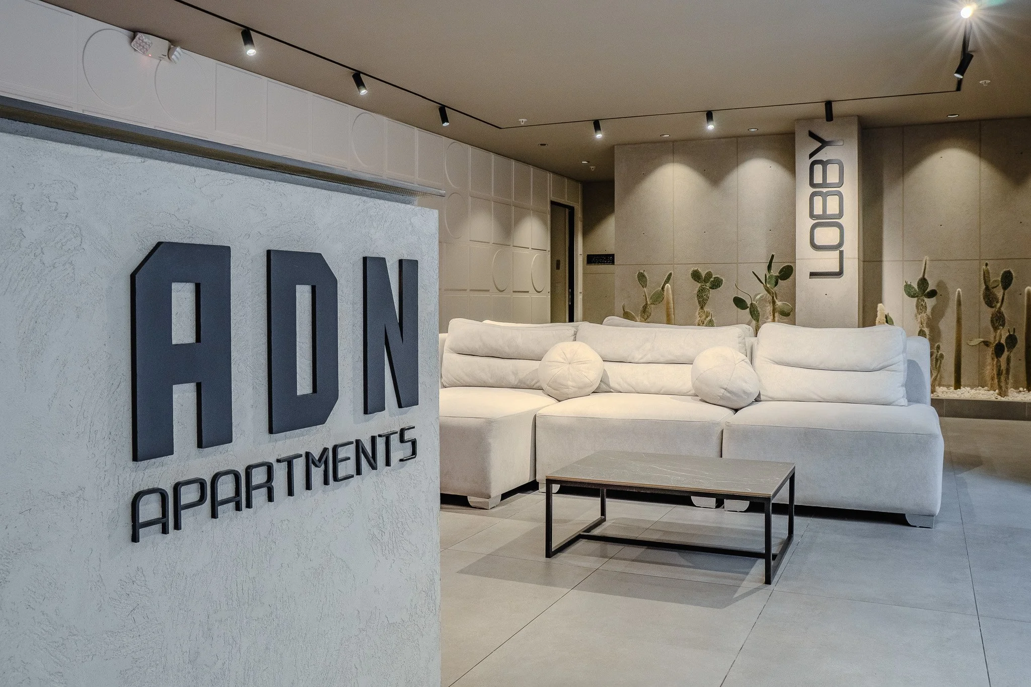Interior lobby of a modern apartment complex with a large white sectional sofa, decorative plants, and a sign that reads "LLOYD B" on a concrete pillar. The background features a wall with the company's logo and a door.