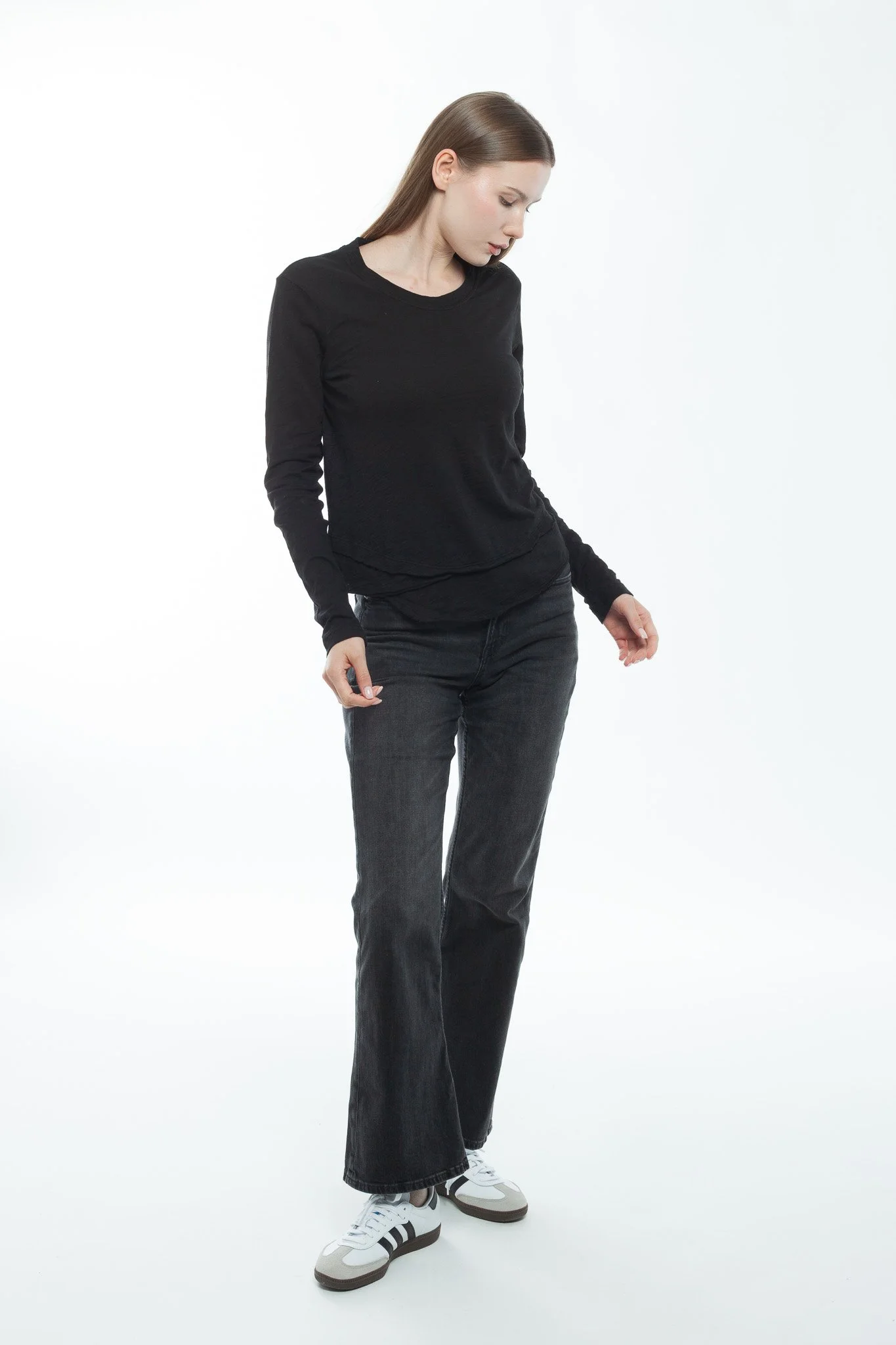 Young woman with long brown hair wearing a black long-sleeve shirt, dark jeans, and white sneakers standing against a white background.