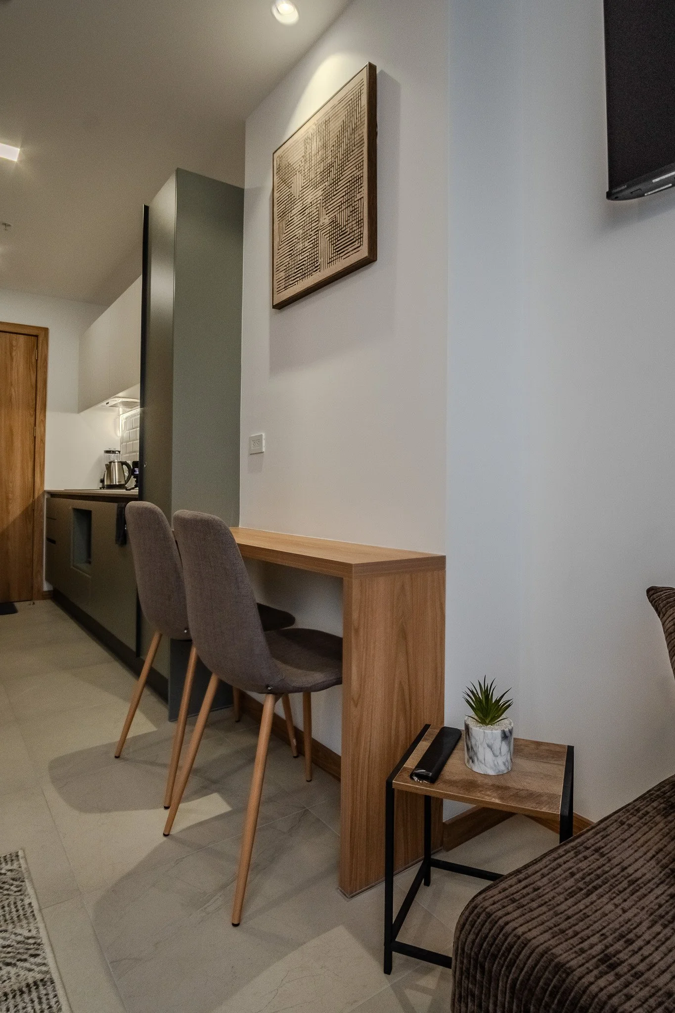 Interior view of a modern living room and dining area with a wooden desk, two gray upholstered chairs, a small side table with a potted plant, and a brown sofa. The walls are light-colored, with a piece of decorative artwork hanging on one wall. The 