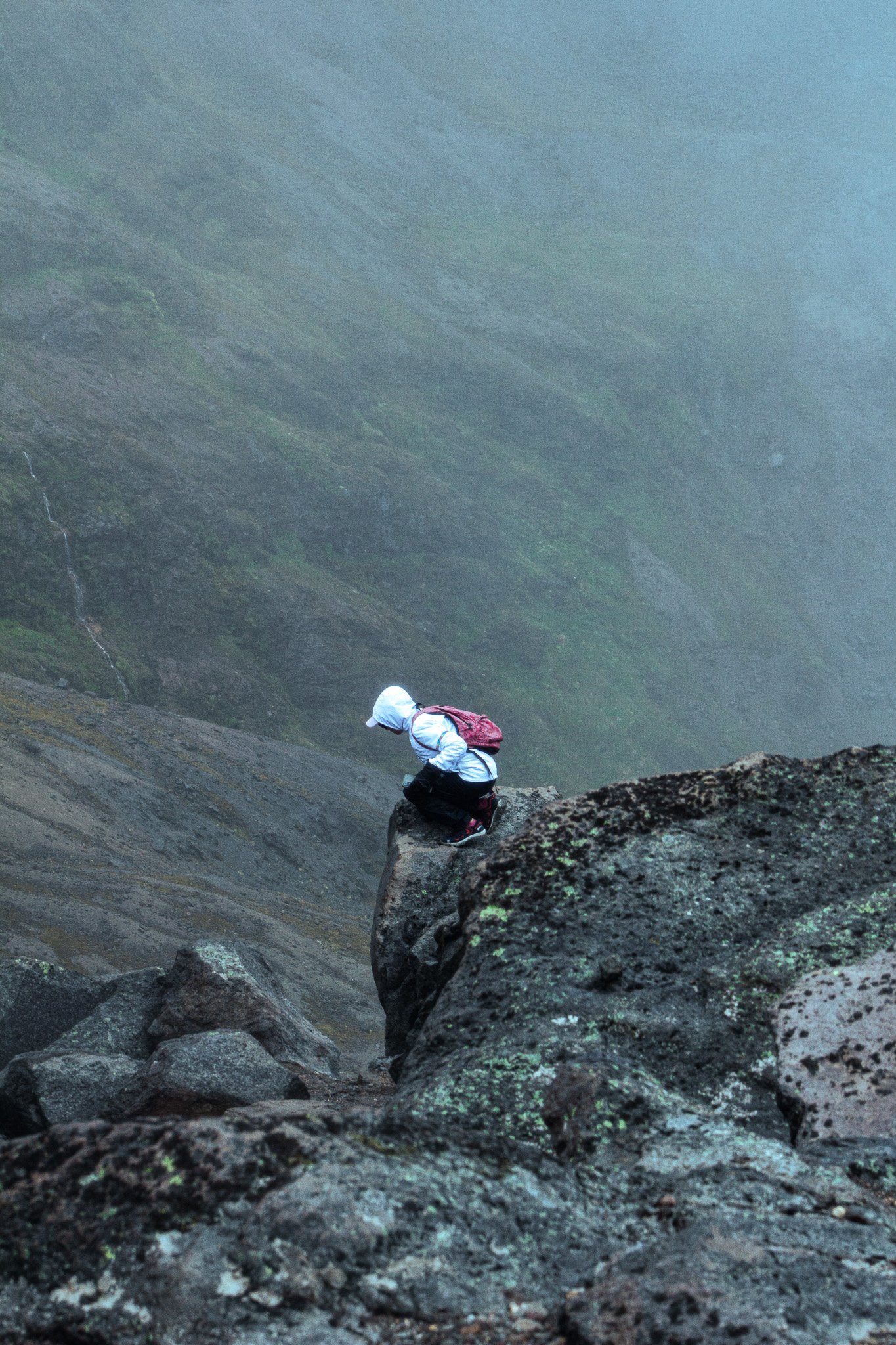 Hiking climber wearing white jacket, black pants, hat, and red backpack sitting on a large rock in a misty, green mountain landscape.