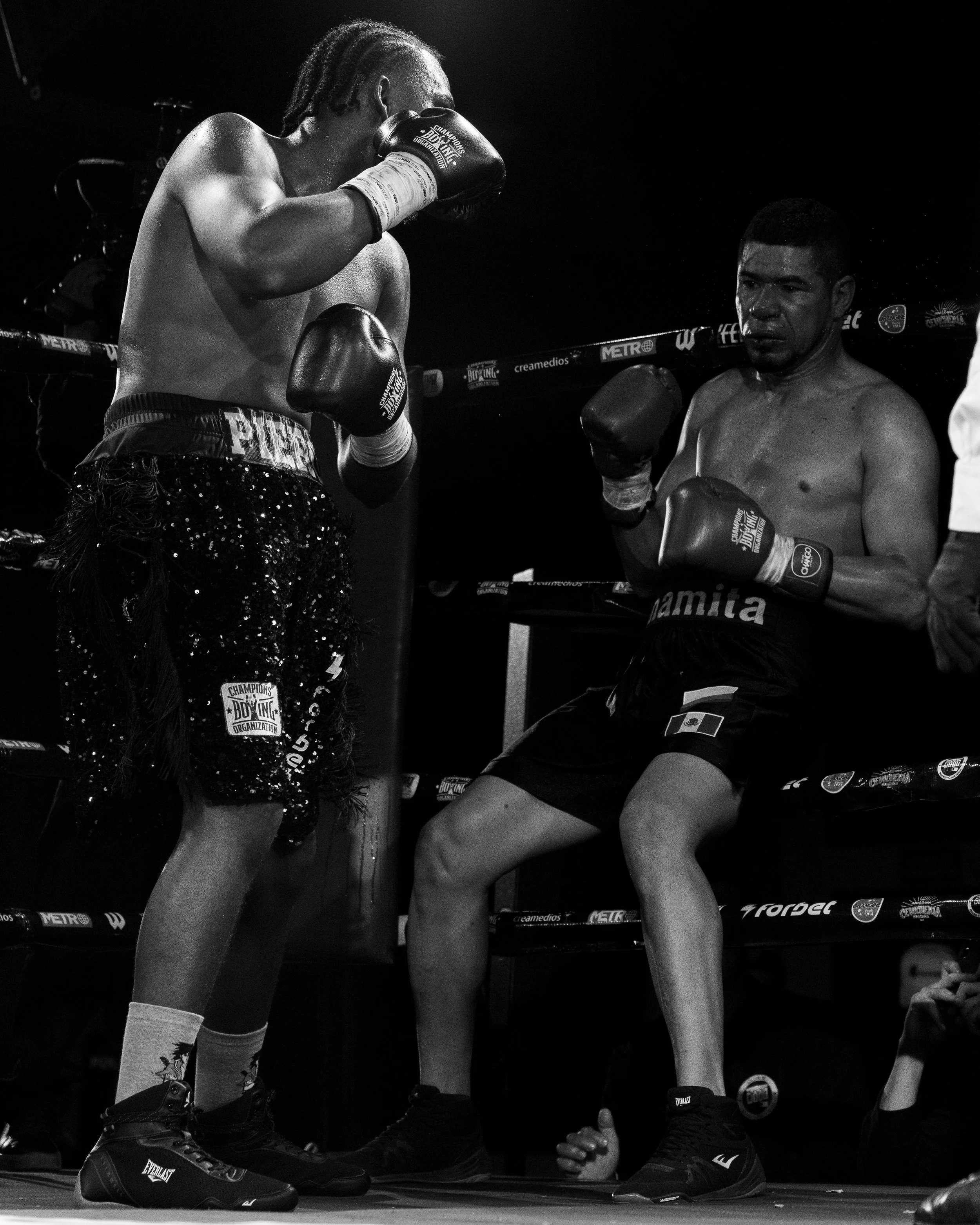 Two male fighters in a boxing ring, in a close-up shot. One is standing with boxing gloves raised, the other is lying on the ground, looking exhausted. Black and white photo.