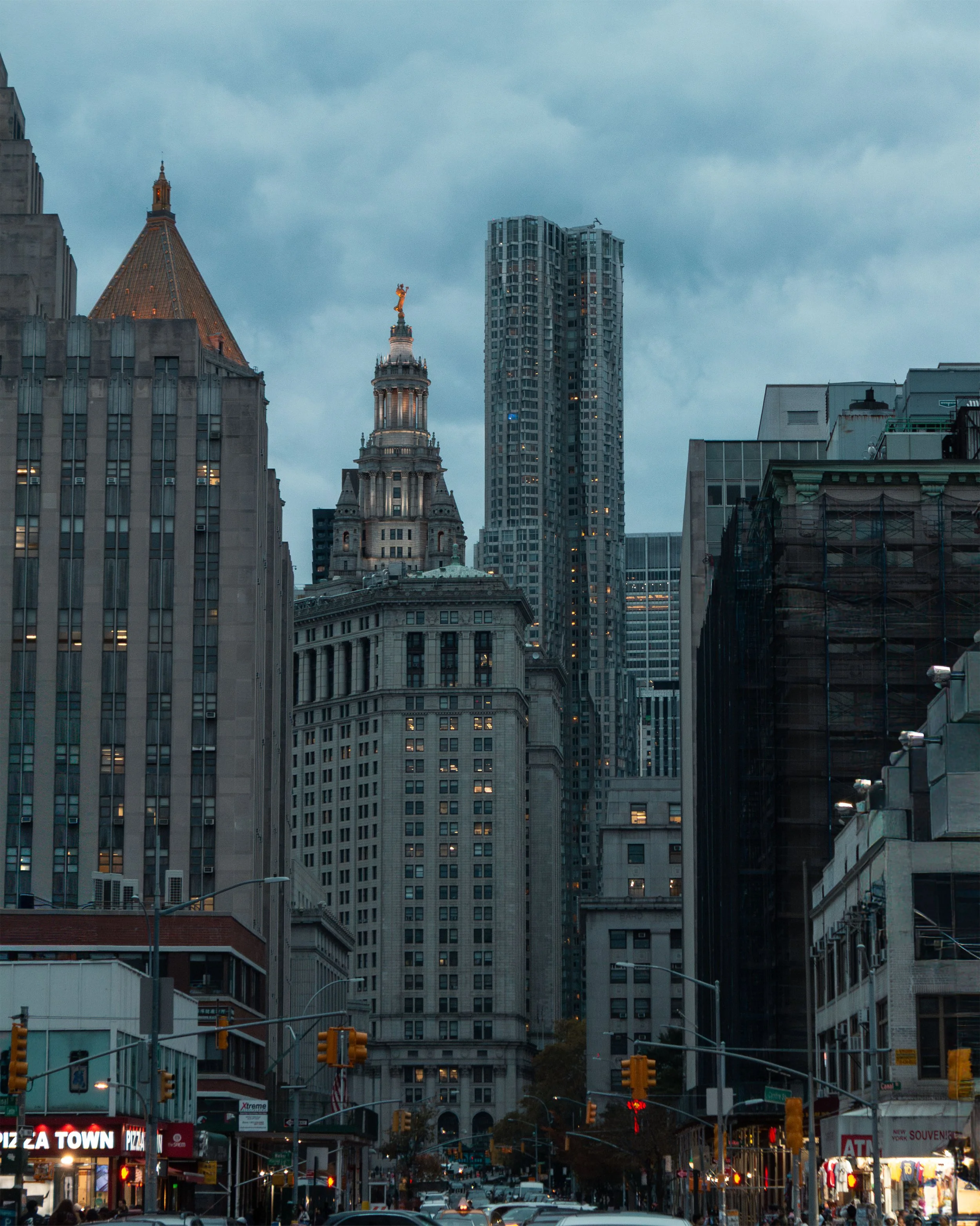Cityscape of skyscrapers with dark clouds overhead, including a historic building with a golden statue on top, and a street with cars and illuminated shops at the bottom.