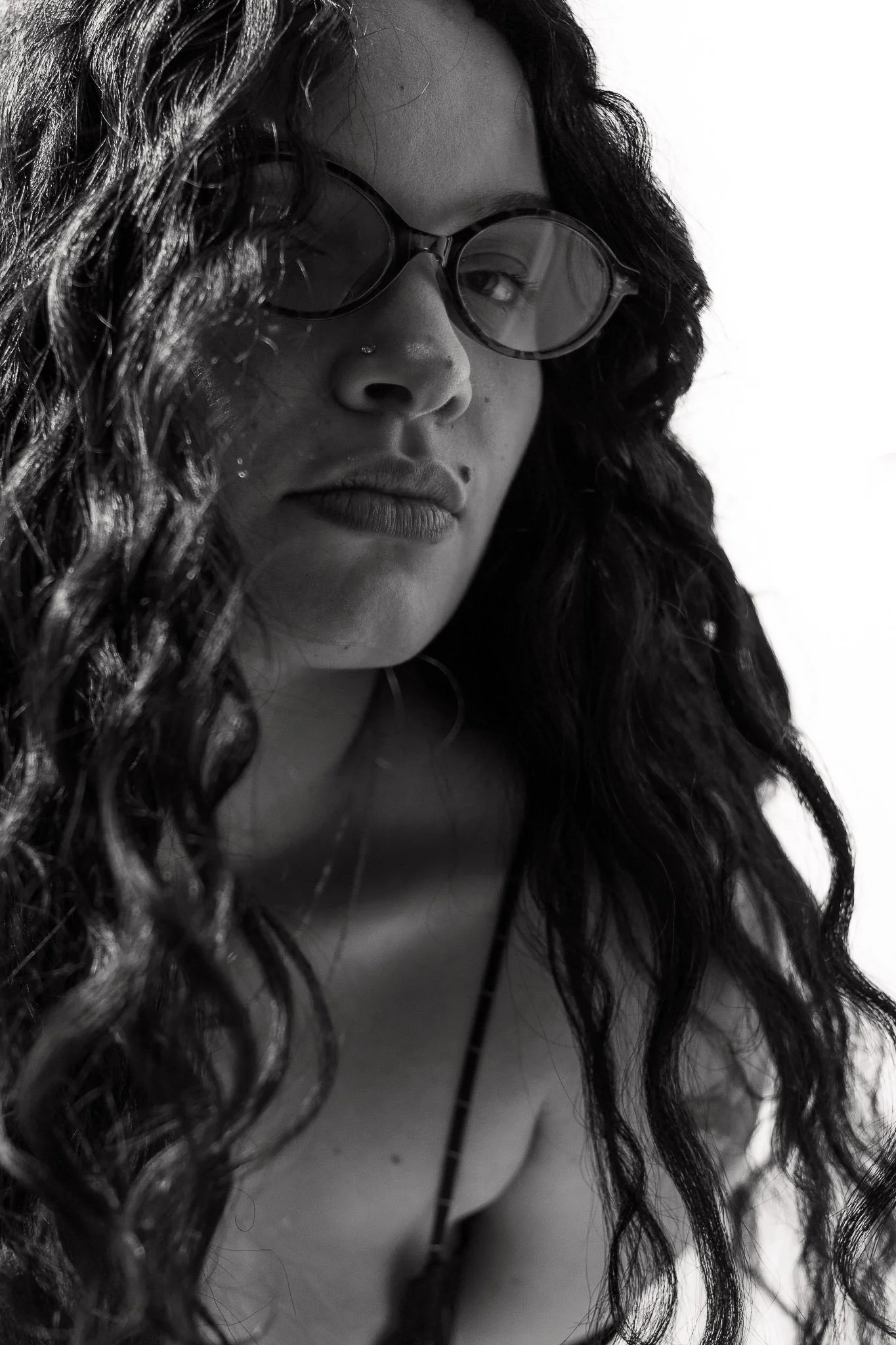 Close-up black and white photo of a woman with long curly hair wearing large dark sunglasses, with a piercing near her nose, looking to the side.