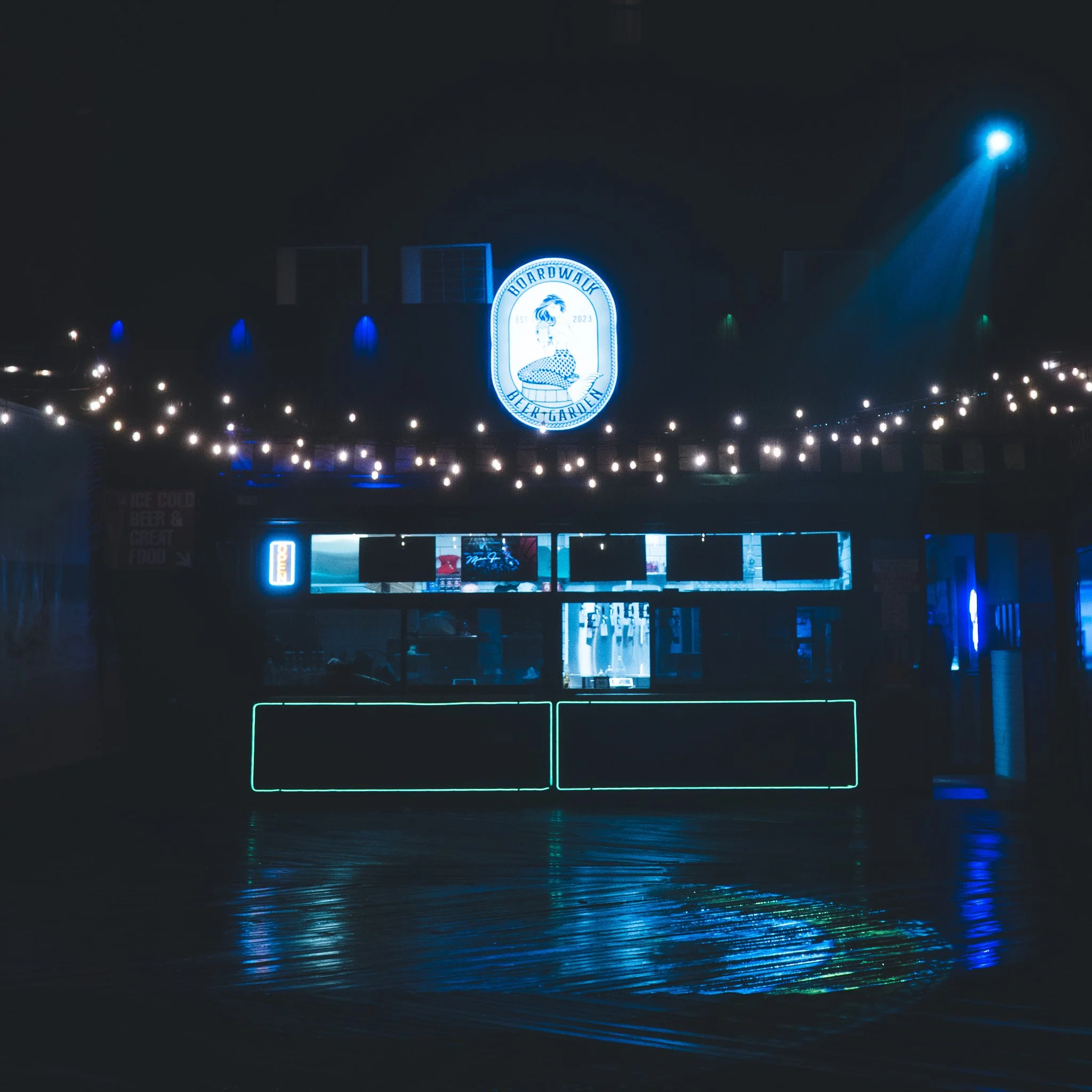 Nighttime scene of a brightly lit sign for Boardwalk Beer Garden with a lion mascot, surrounded by dark sky and reflected lights on a wet surface.
