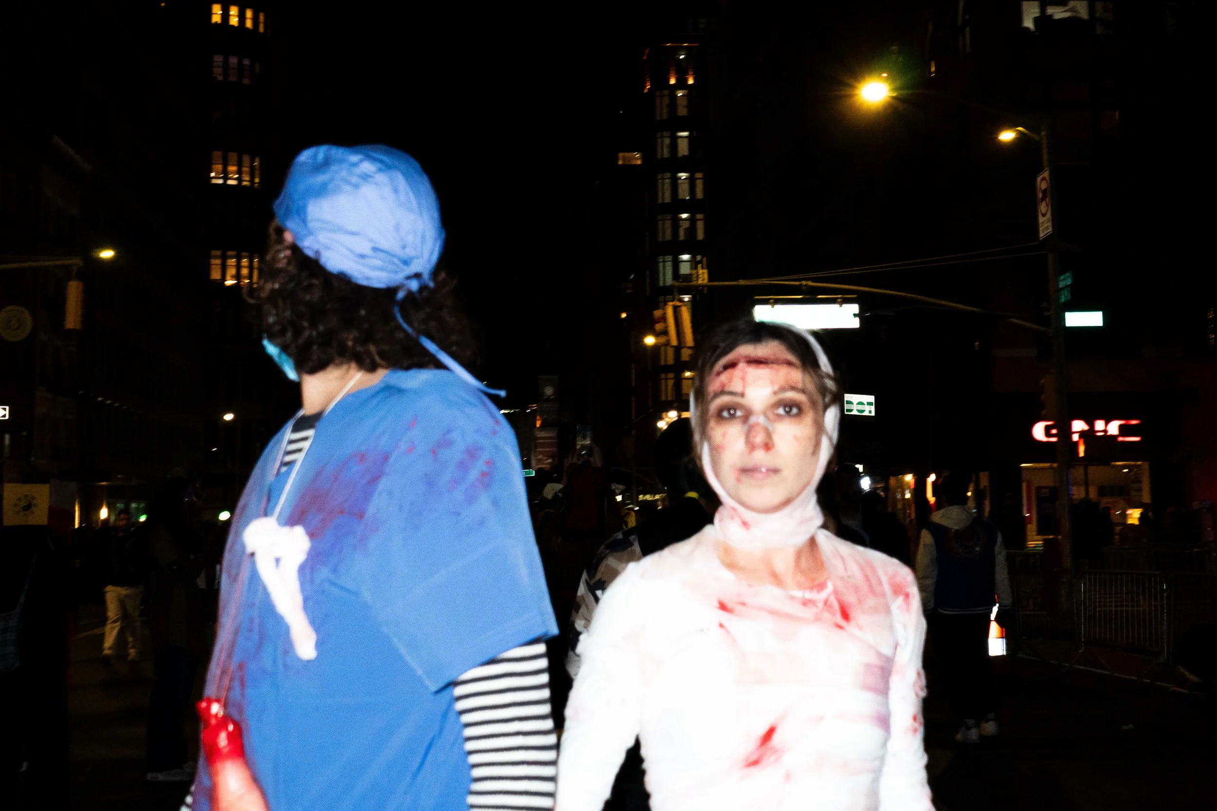 Two people dressed as injured medical personnel at night, one with a blue surgical gown and cap, the other with a bloodstained white outfit and head bandage, on a city street.