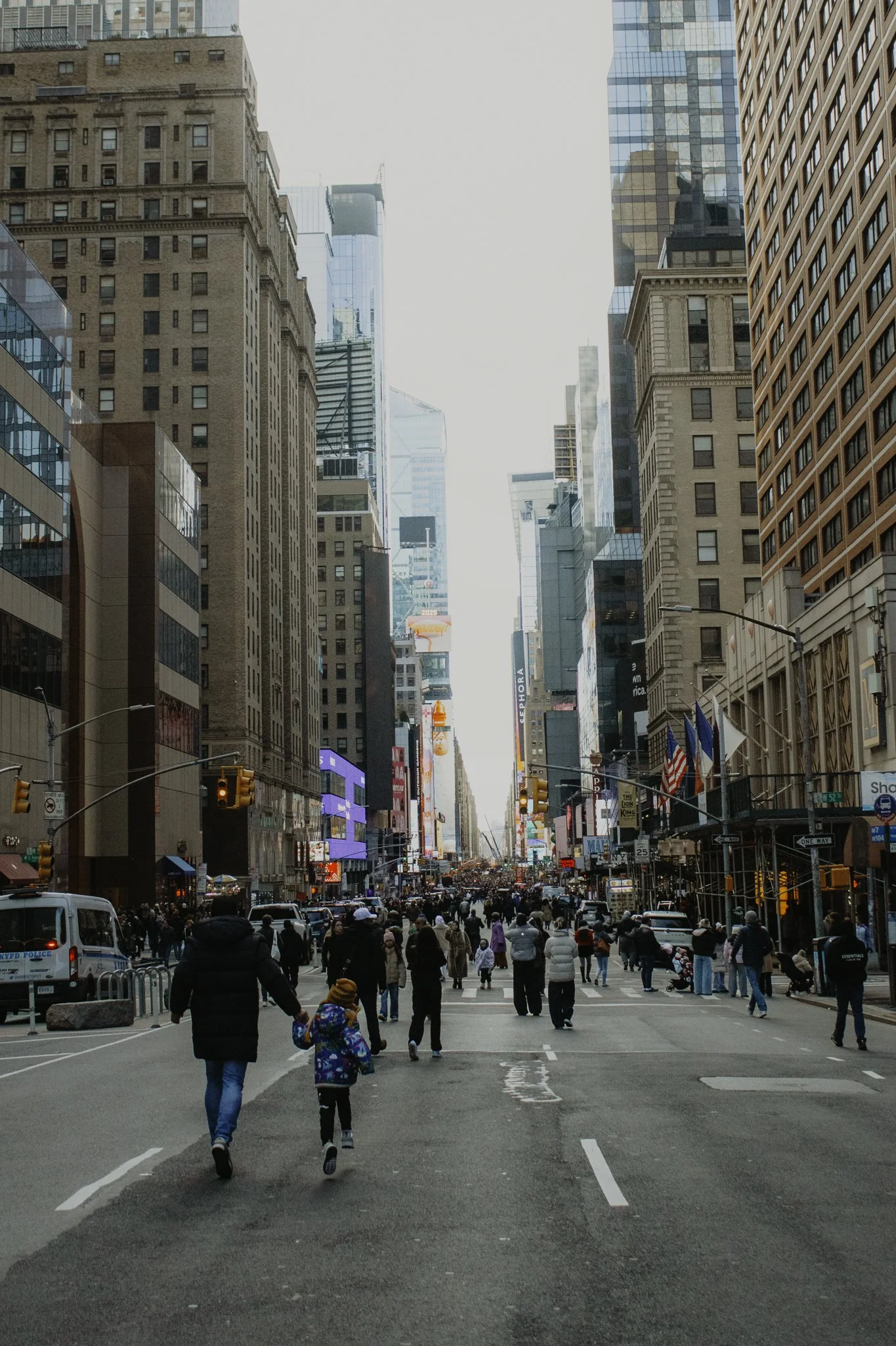 A crowded city street scene with many pedestrians walking across a crosswalk, tall buildings on both sides, and electronic billboards and flags along the street.