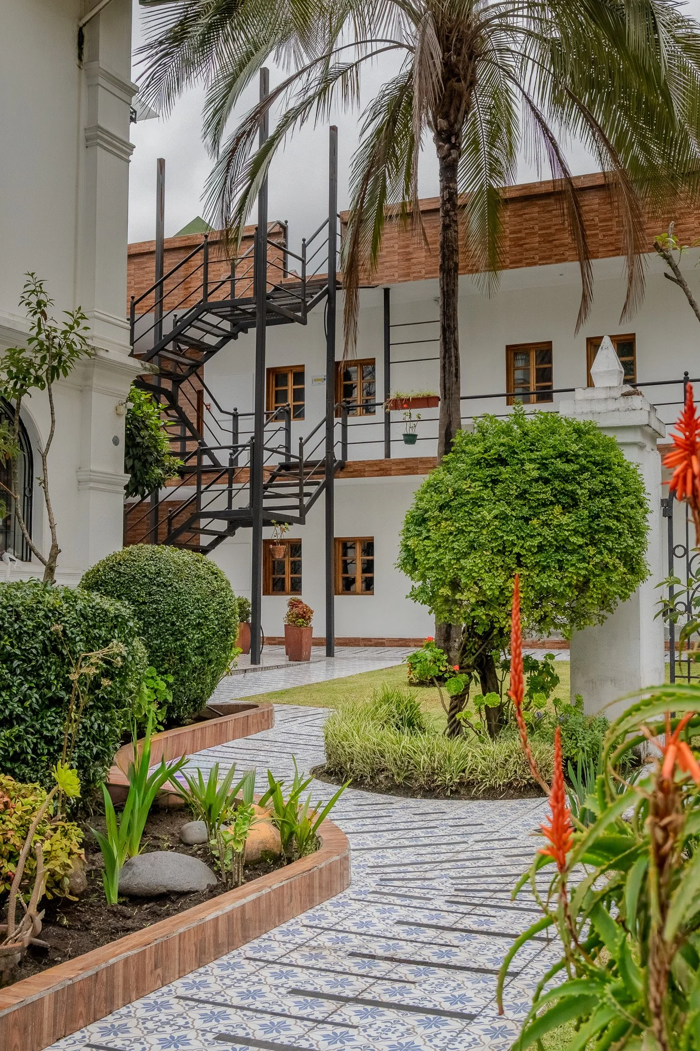 A courtyard garden with potted plants, trimmed bushes, and a tiled pathway. A black metal fire escape staircase leads to the second floor of a white building with wooden window frames. There are palm trees and flowering plants, and the sky is overcas