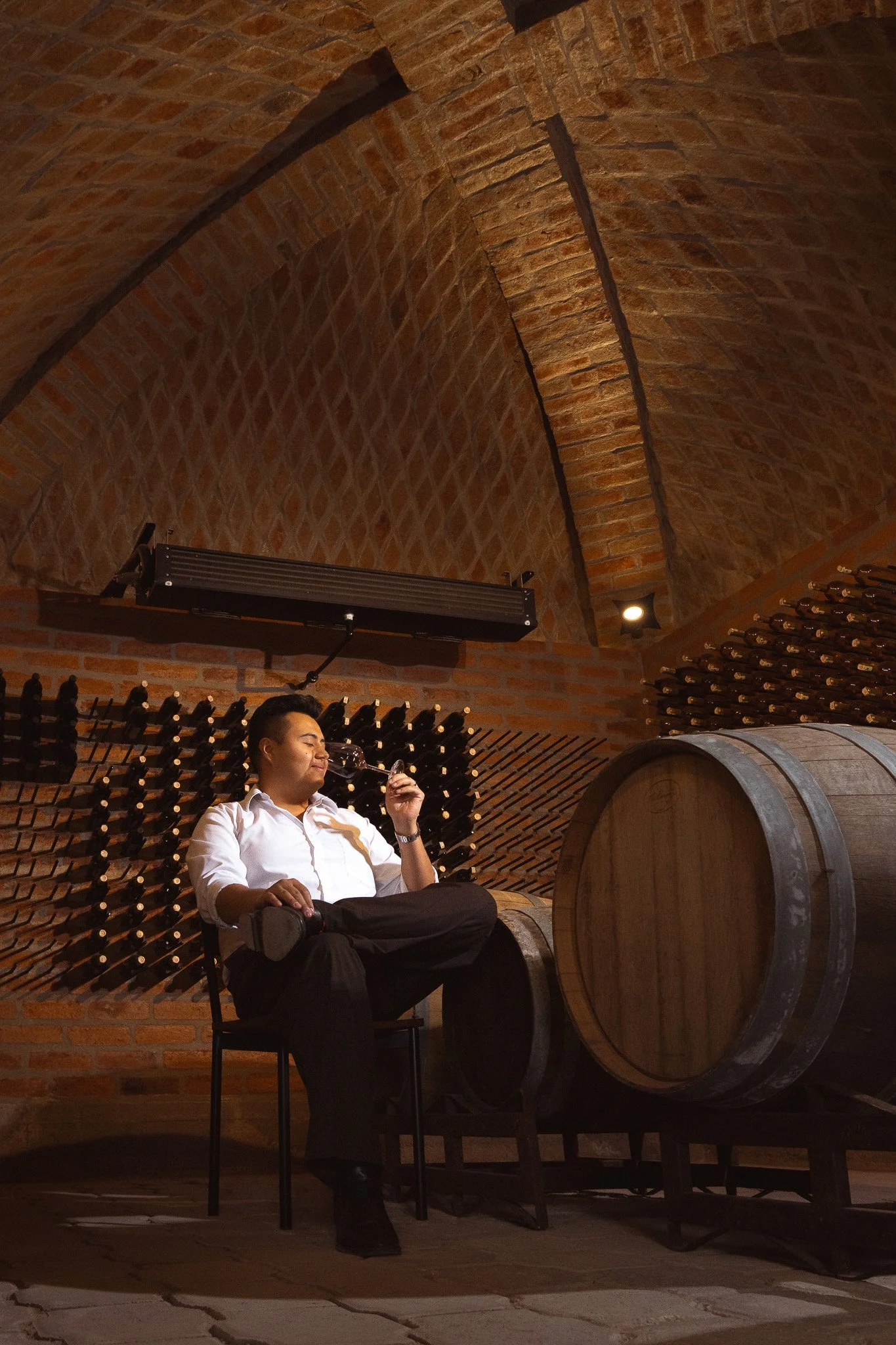 A man in a white shirt and black pants sitting on a chair with his eyes closed, holding a glass of wine, inside a wine cellar or tasting room with brick vaulted ceiling, wine racks, and a large wine barrel nearby.