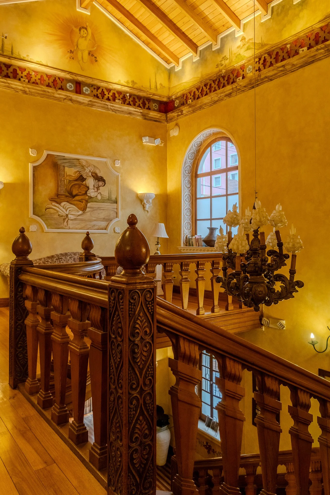 Inside a house, a staircase with wooden balustrades and newel posts leading down to a lower level. The wall features a large portrait of a woman sitting, and a decorative chandelier hanging from the ceiling. There are arched windows allowing natural 