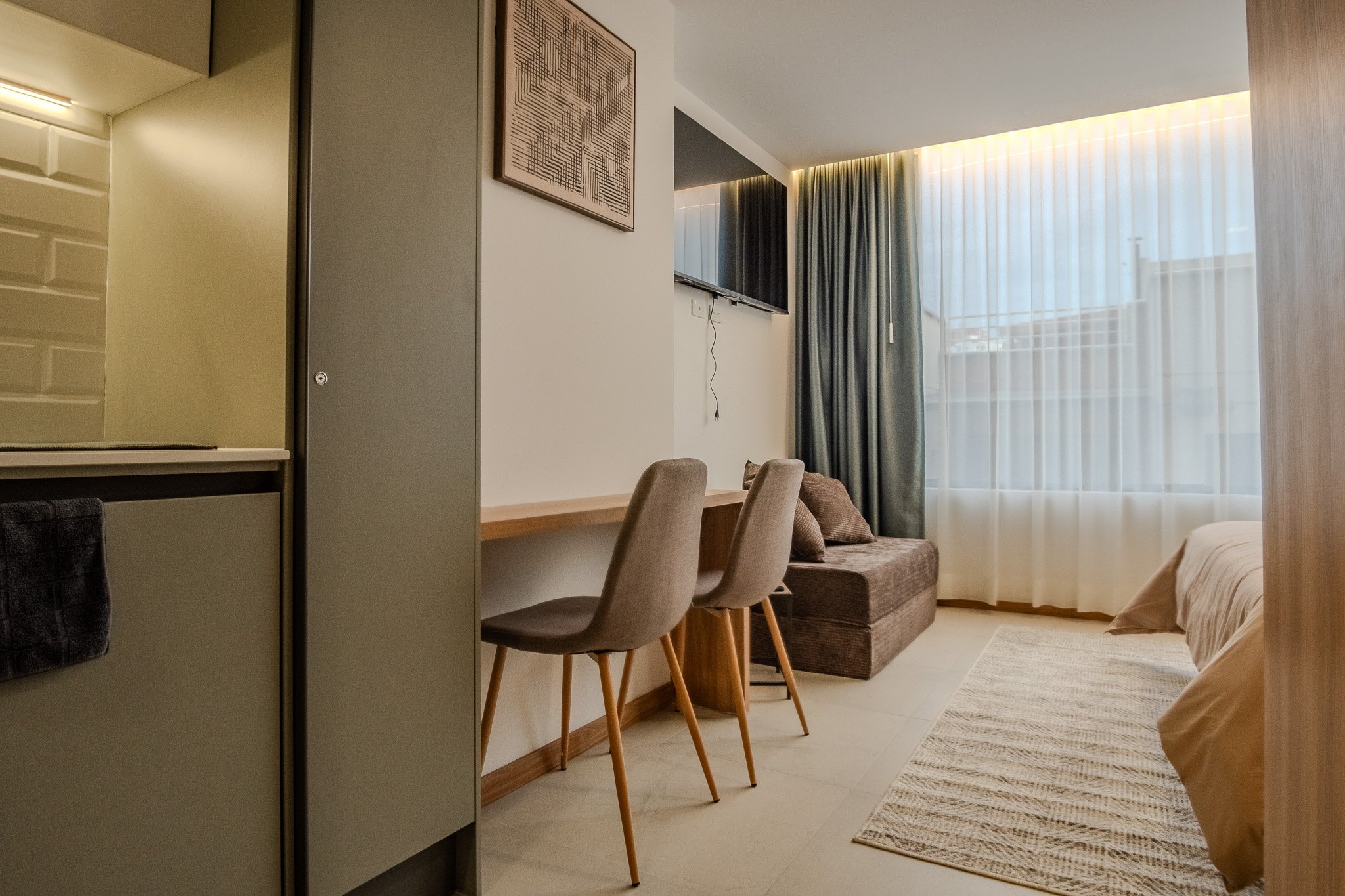 Interior of a modern hotel room with a bed, a sitting area with chairs, a desk, wall-mounted TV, curtains, and a textured white wall.
