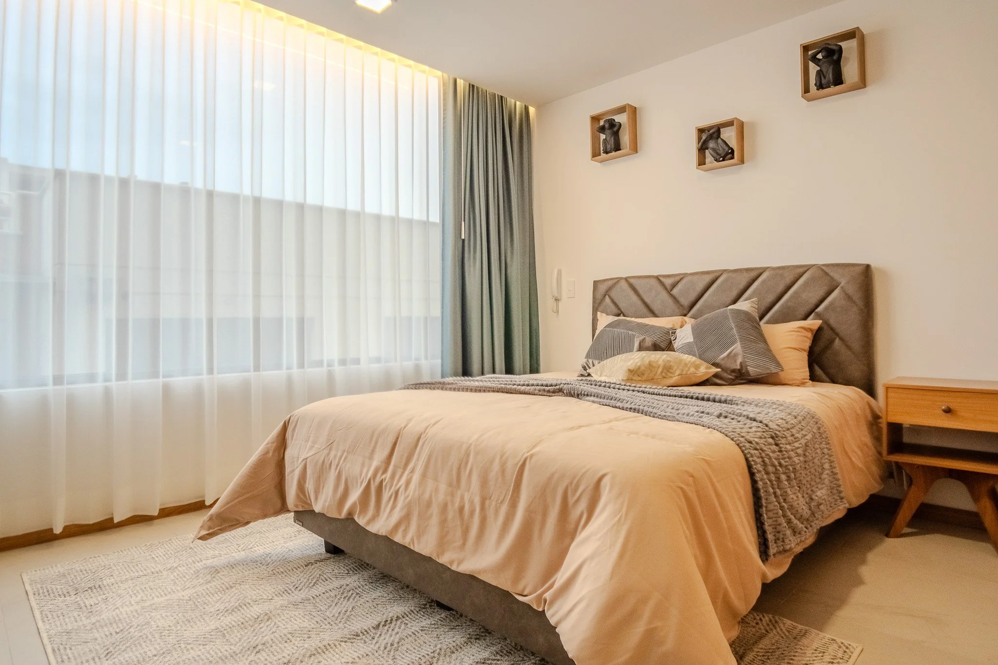 A cozy bedroom with beige bedding, a tufted headboard, and decorative pillows. A wooden nightstand is beside the bed, and wall-mounted shelves with small sculptures are above. Sheer curtains cover large windows, letting in natural light.