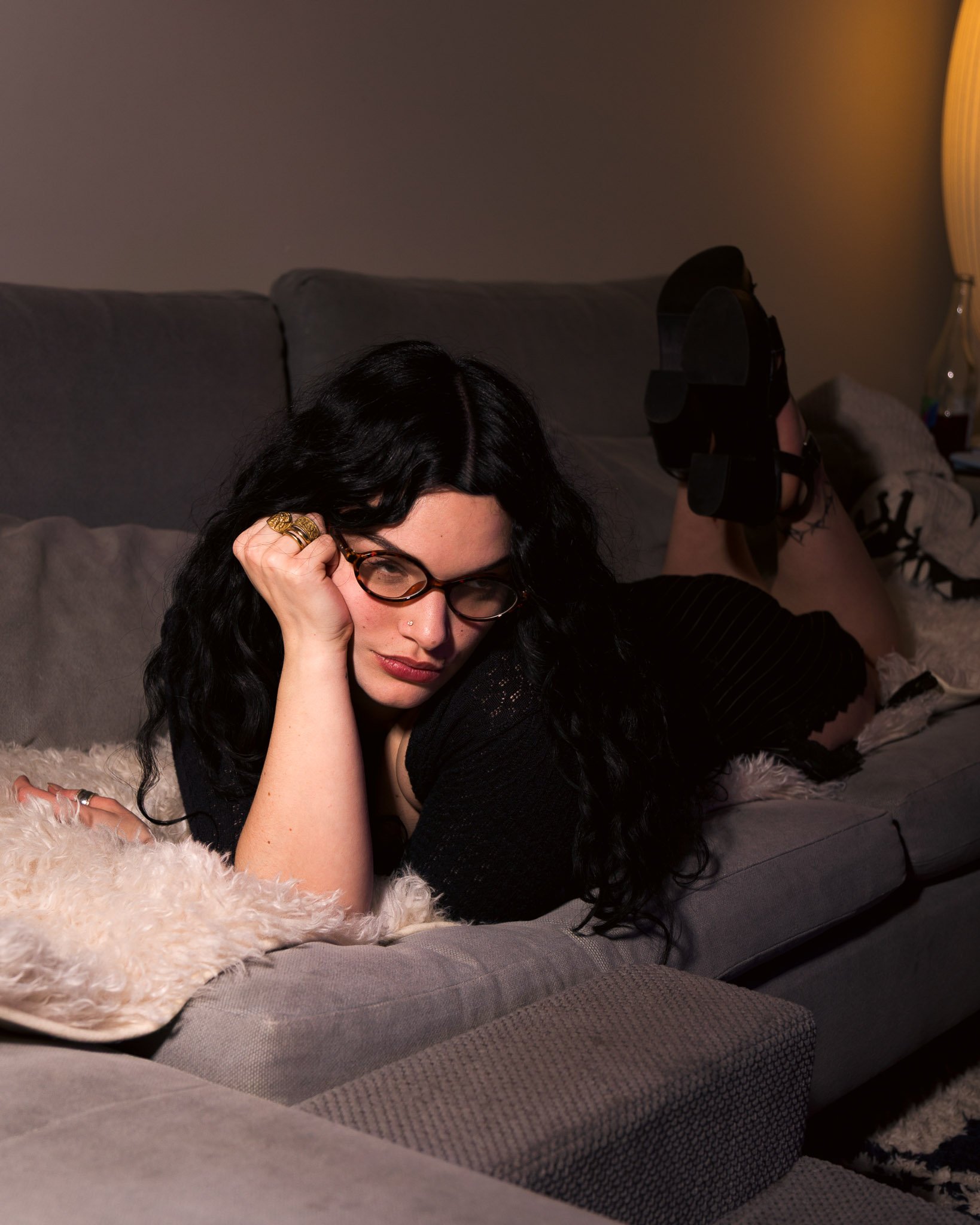 A woman with black hair and glasses, lying on a gray couch with a furry blanket, resting her head on her hand, and wearing multiple rings. She appears relaxed in a cozy living room setting, with part of her face and one shoe visible in the image.