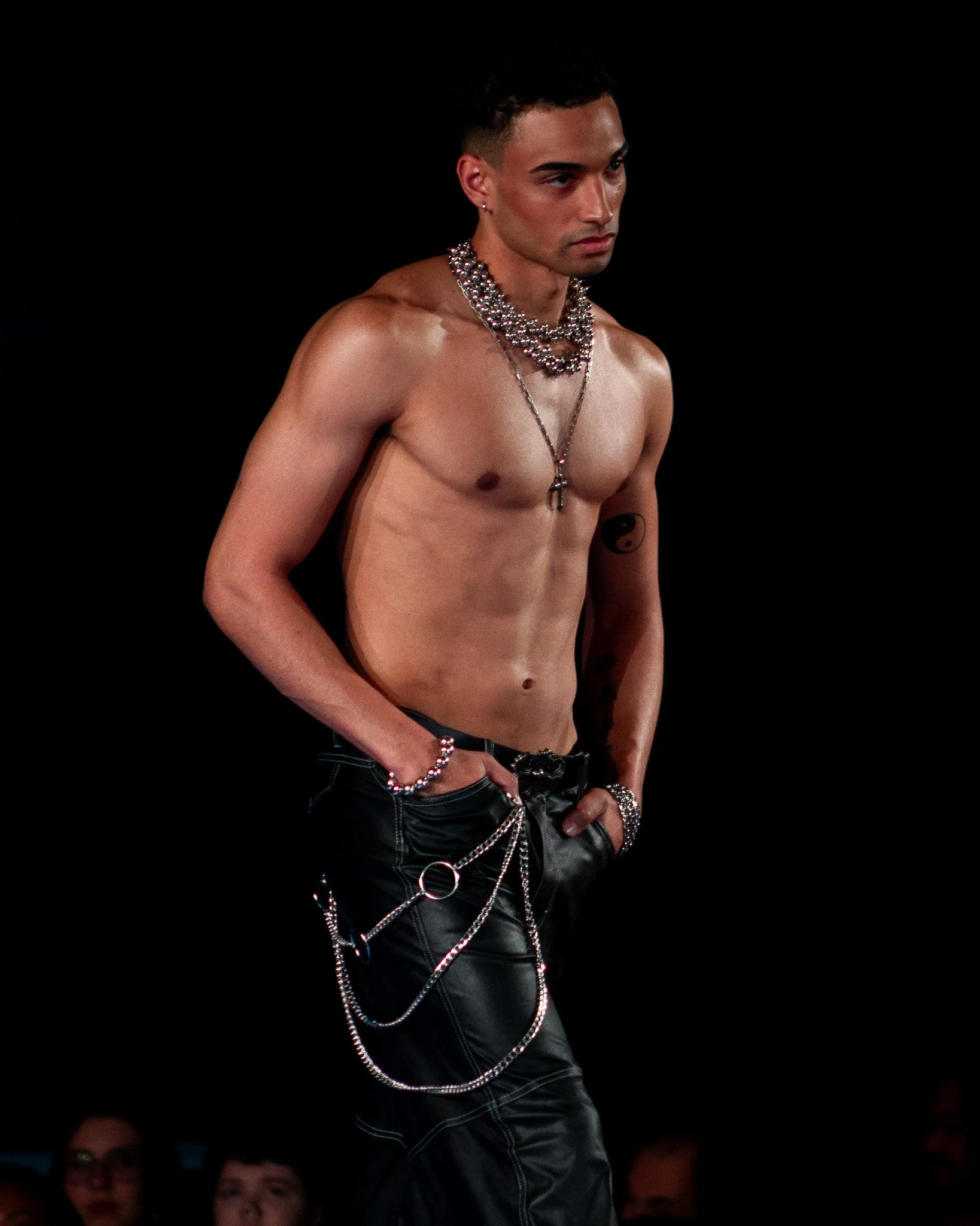 A shirtless man wearing layered necklaces, chains, rings, and bracelets stands against a black background, with an audience observing in the lower part of the image.