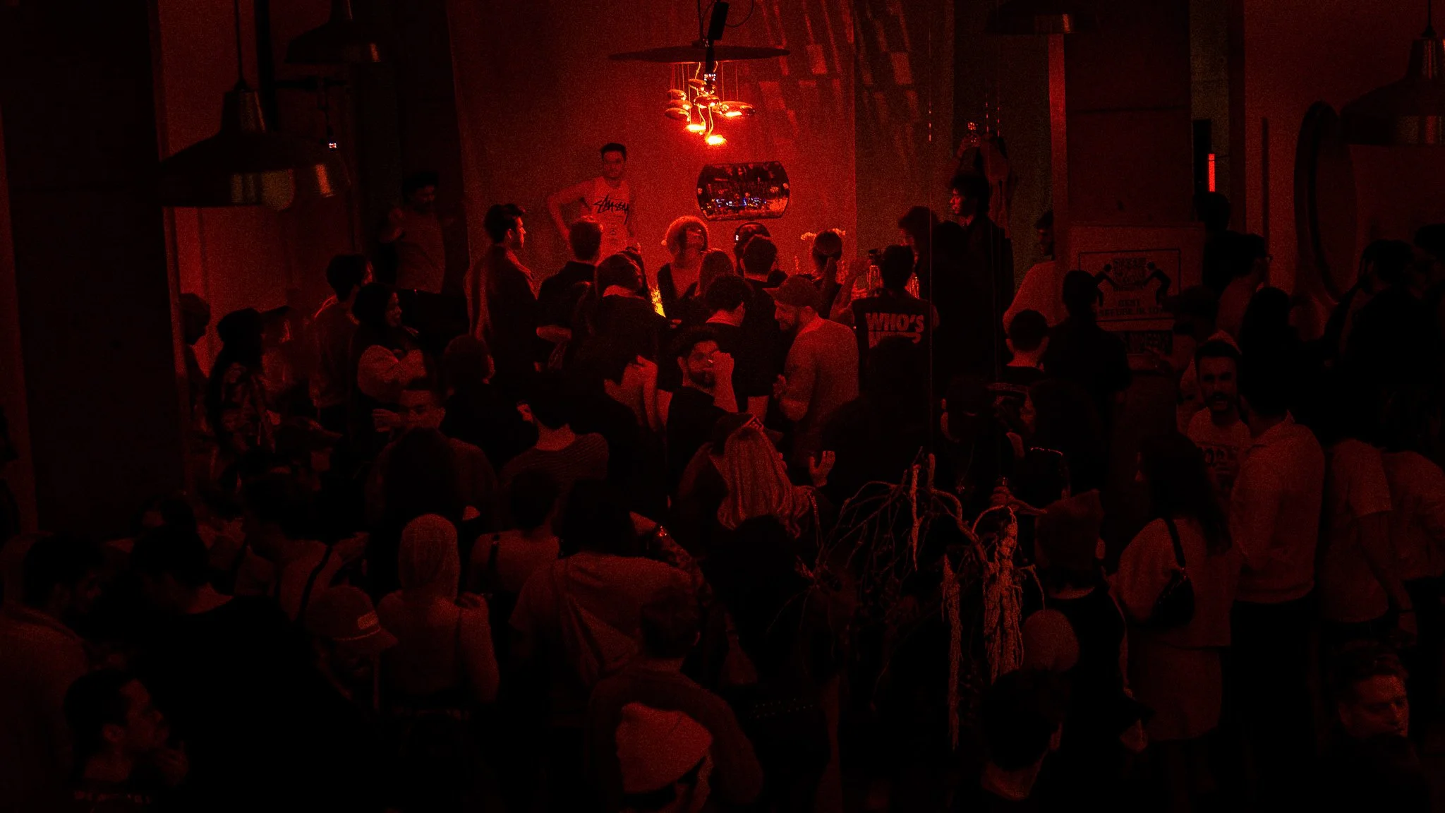 A crowded indoor party or gathering with people socializing in dim red lighting, with a chandelier and some decorative elements visible in the background.
