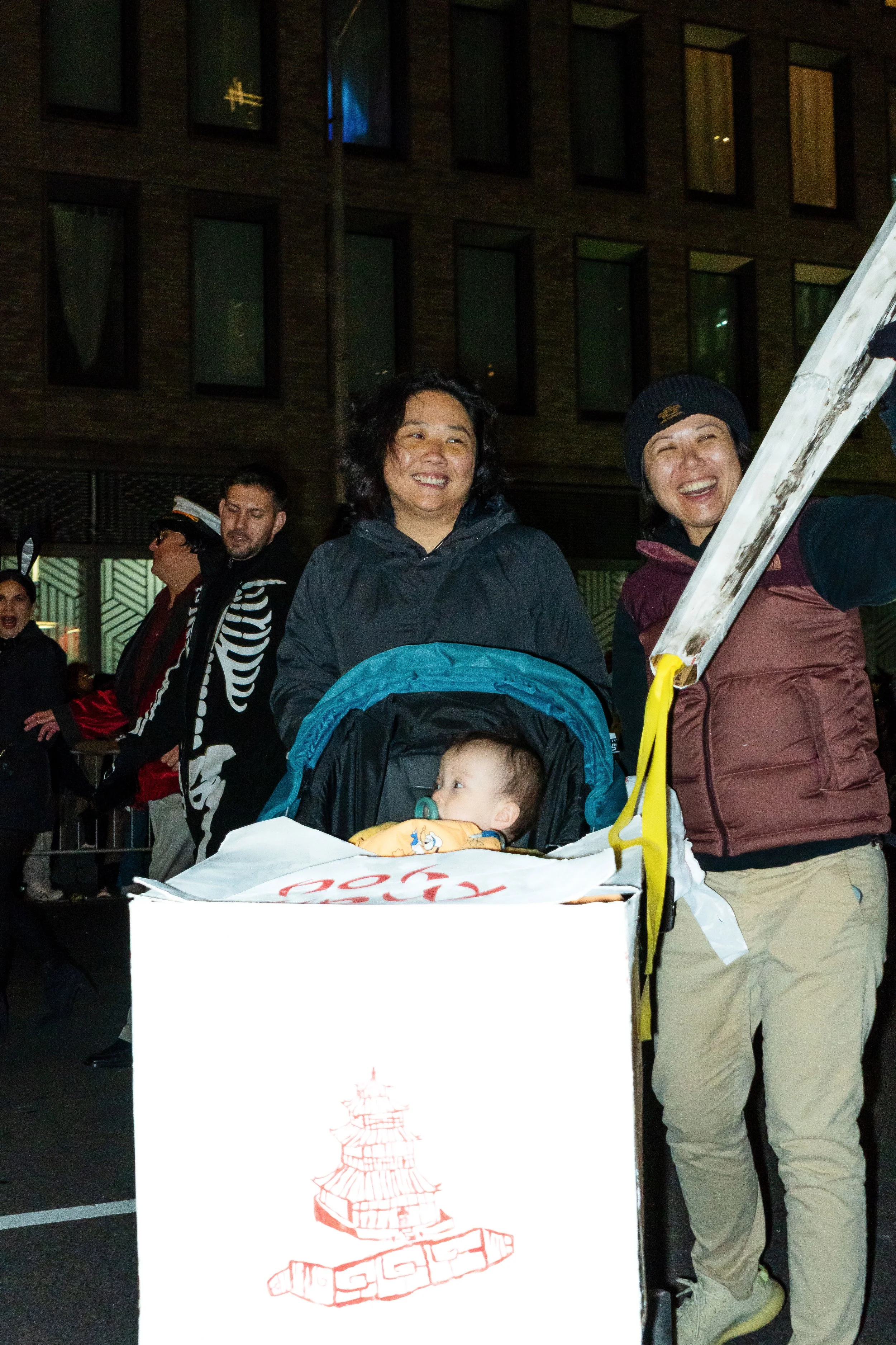 People gathered at night, some smiling, with a woman pushing a stroller with a baby inside, in an urban setting.