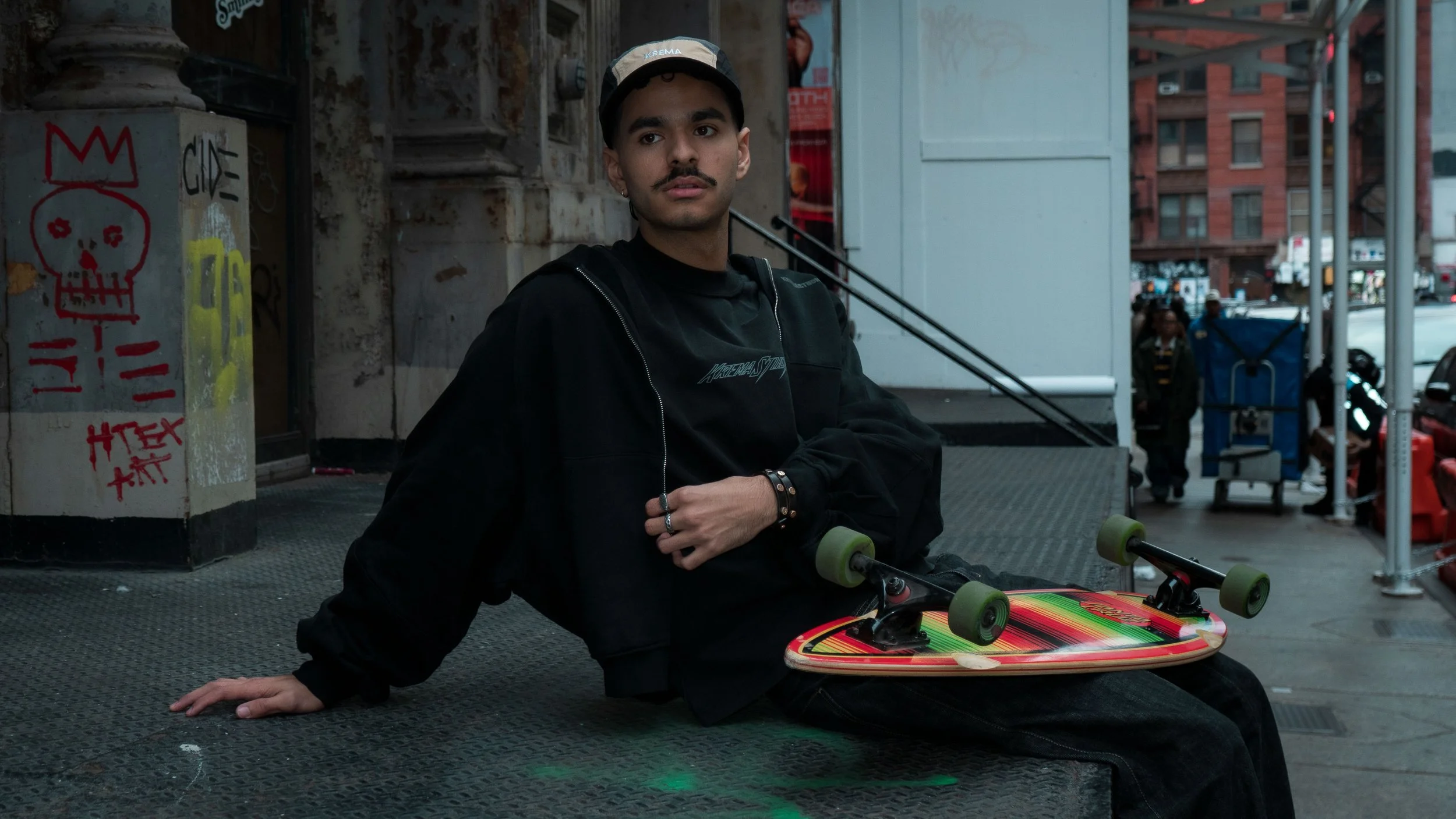 Young man with a mustache and black hair wearing a beige cap, black jacket, and black shirt sitting on a city sidewalk with a skateboard on his lap, in an urban environment with graffiti and buildings in the background.