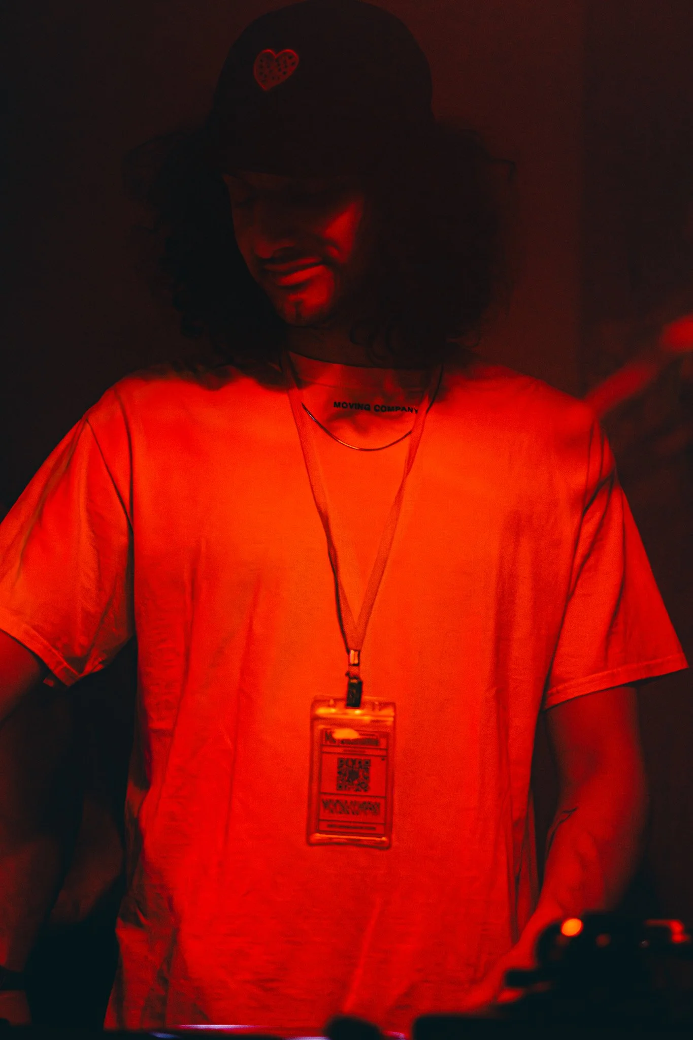 A person wearing a cap with a red heart and a shirt with the words "MOVING COMPANY" on the neck, illuminated in red light, with a lanyard and an ID badge around their neck, in a dark environment.